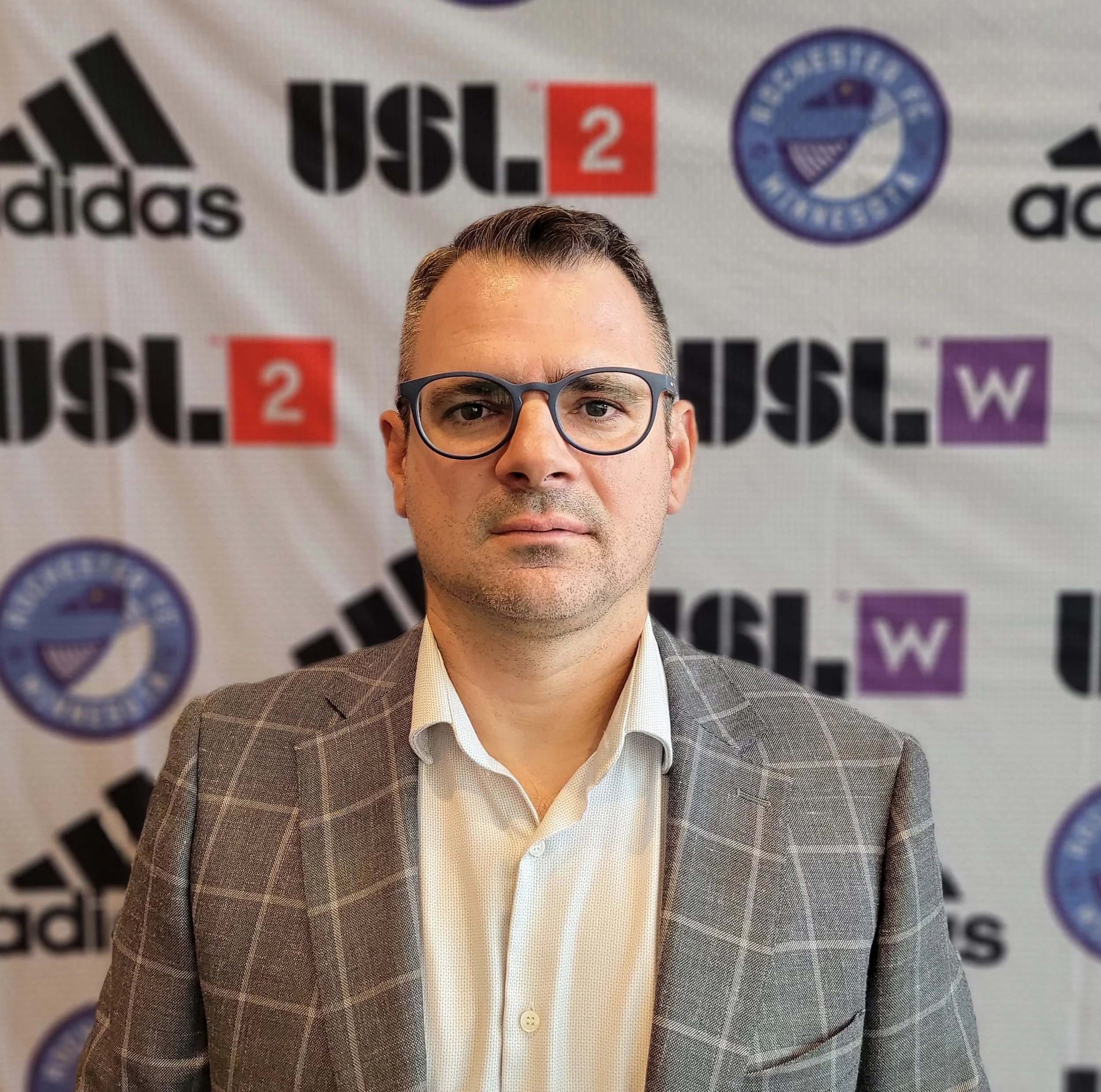 Close-up of a man in glasses and a checked blazer, with a backdrop featuring logos including Adidas, USL 2, and a blue circular emblem.