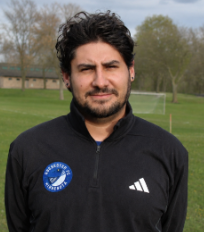 Portrait of a man with dark curly hair and a beard, wearing a black jacket with a sports logo, standing outdoors in a park with trees and a field.