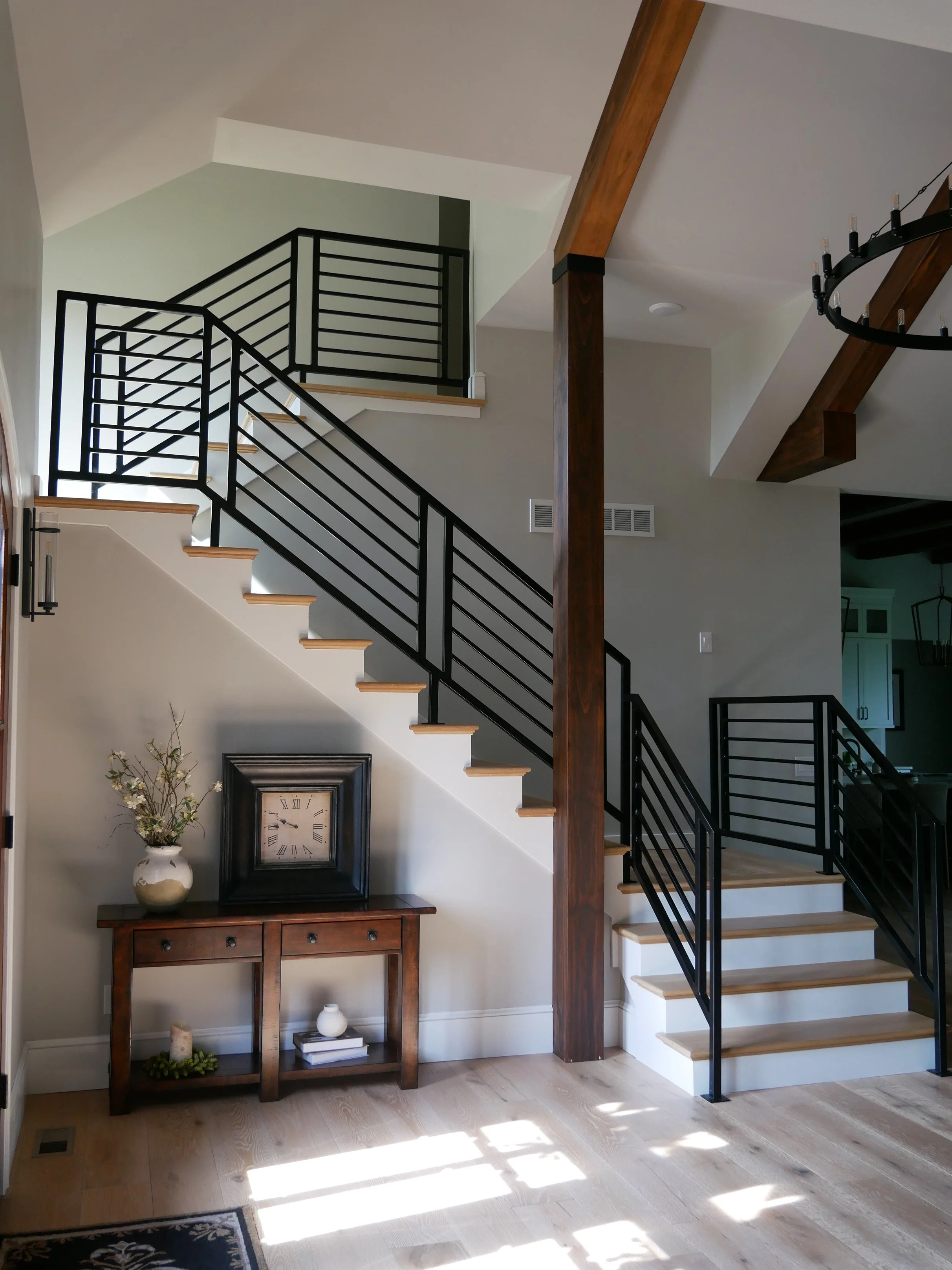 Interior view of a staircase with black metal railings and wooden steps in a modern home. A wooden pillar supports the ceiling, and a small wooden console table with decor items stands against the wall below. Natural light casts shadows on the wooden