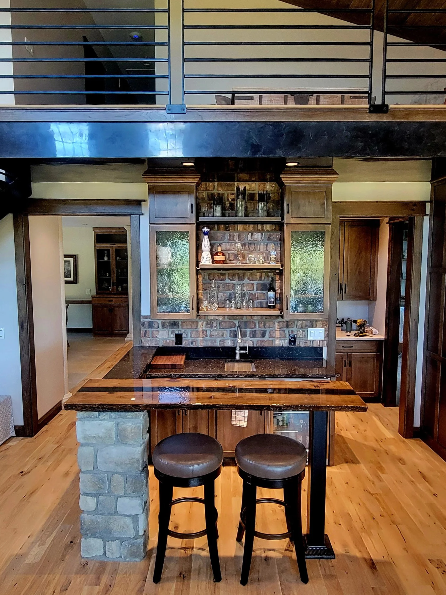 A kitchen with a black granite countertop, brick wall, and wooden cabinets. Two bar stools are in front of the counter, with a breakfast bar made of wood and black granite. In the background, a doorway leads to another room, and there are decorative 