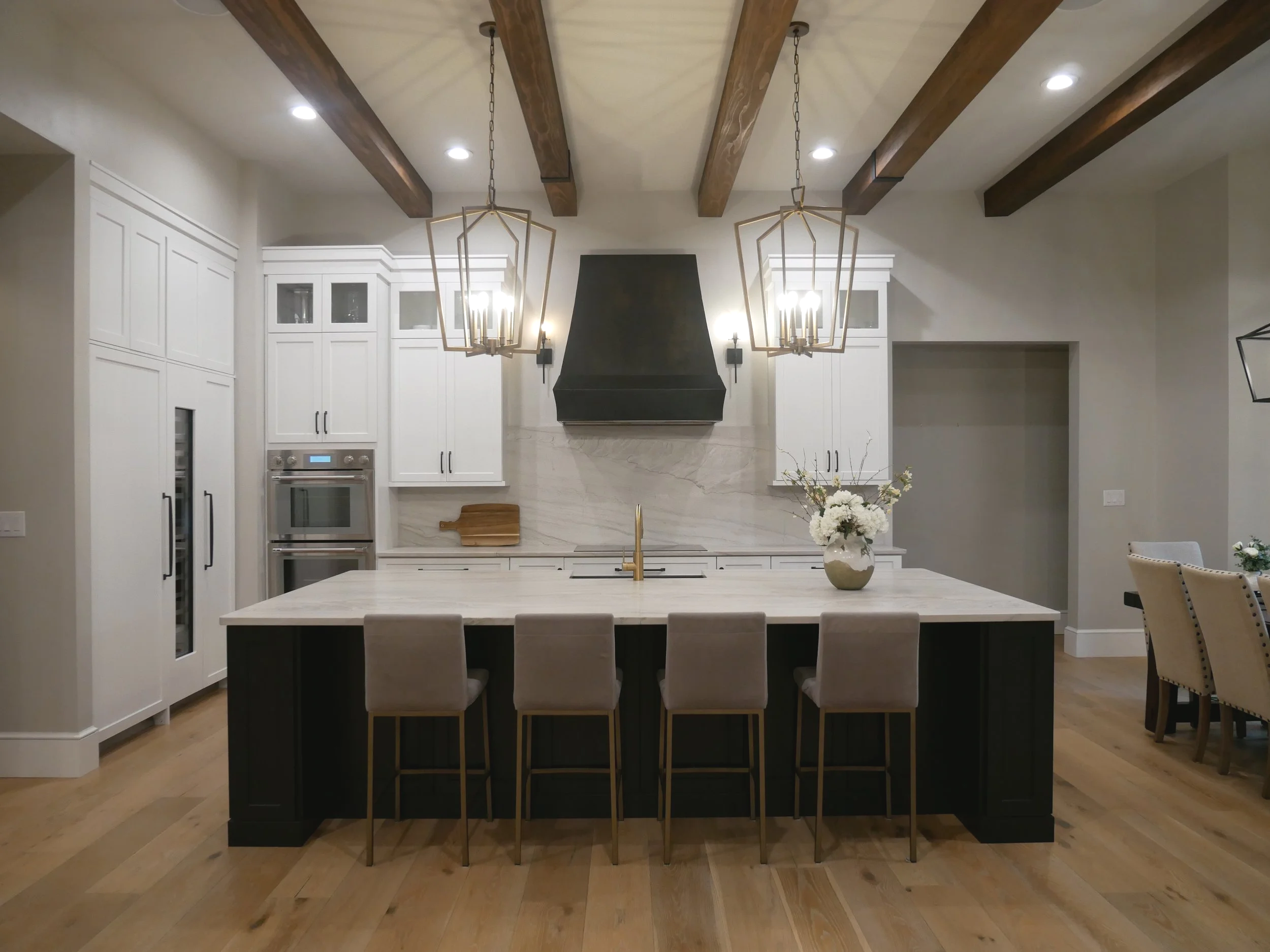 Modern kitchen with white cabinets, black island, two hanging geometric light fixtures, stainless steel oven, black range hood, marble backsplash, and a vase of white flowers on the island.