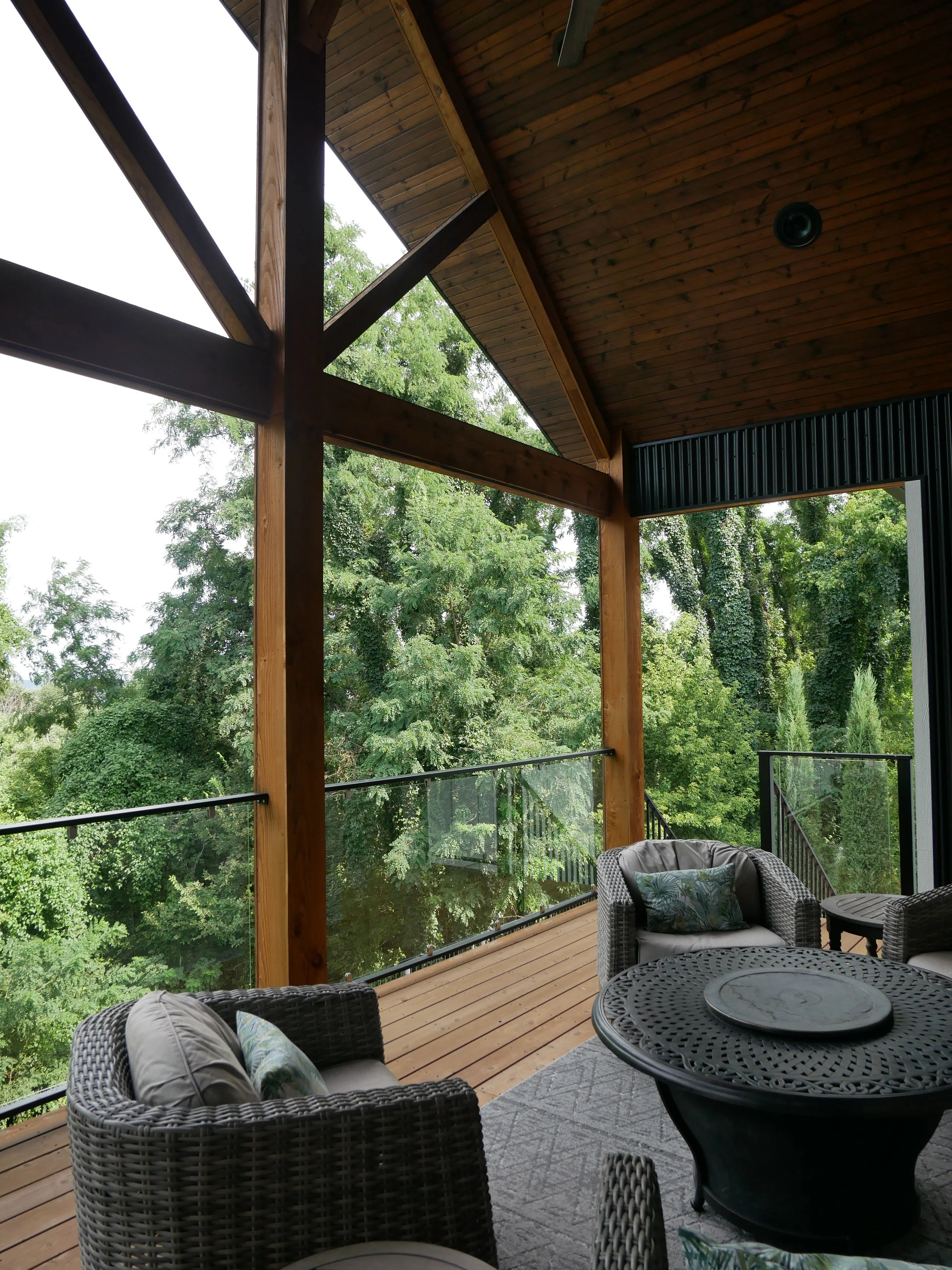A covered outdoor balcony with wicker chairs, a glass-top table, and lush green trees in the background.