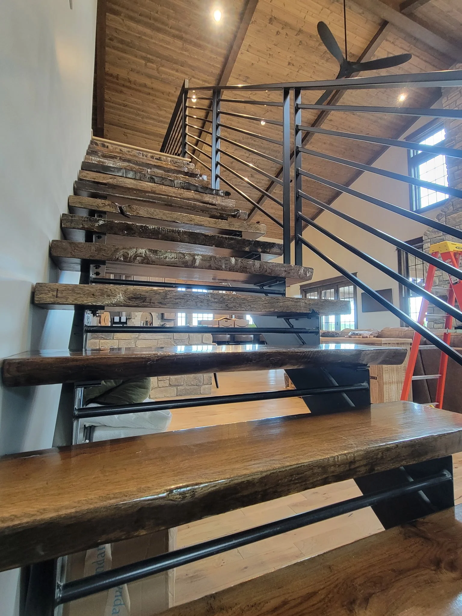 A staircase with thick wooden steps and black metal railings inside a room with wooden ceiling panels and brick walls. A ceiling fan is visible near the ceiling.
