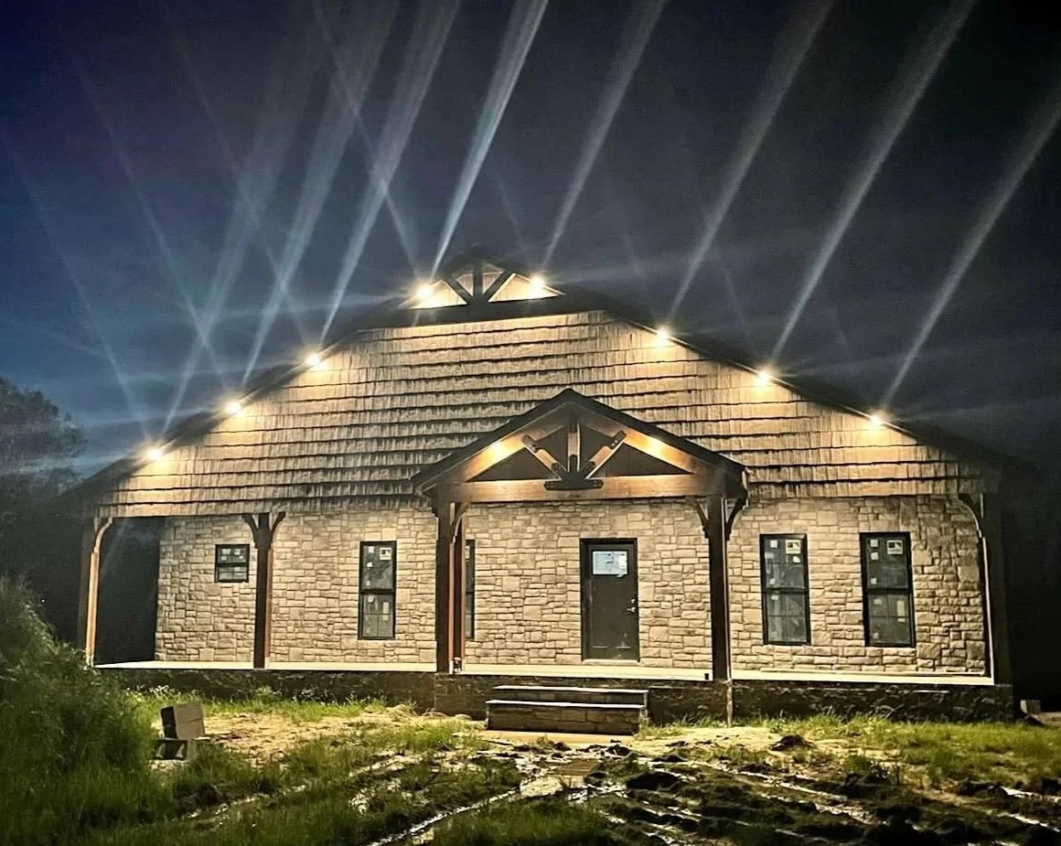 A house at night illuminated by exterior lights, with stone walls, a gabled roof, and wooden accents.