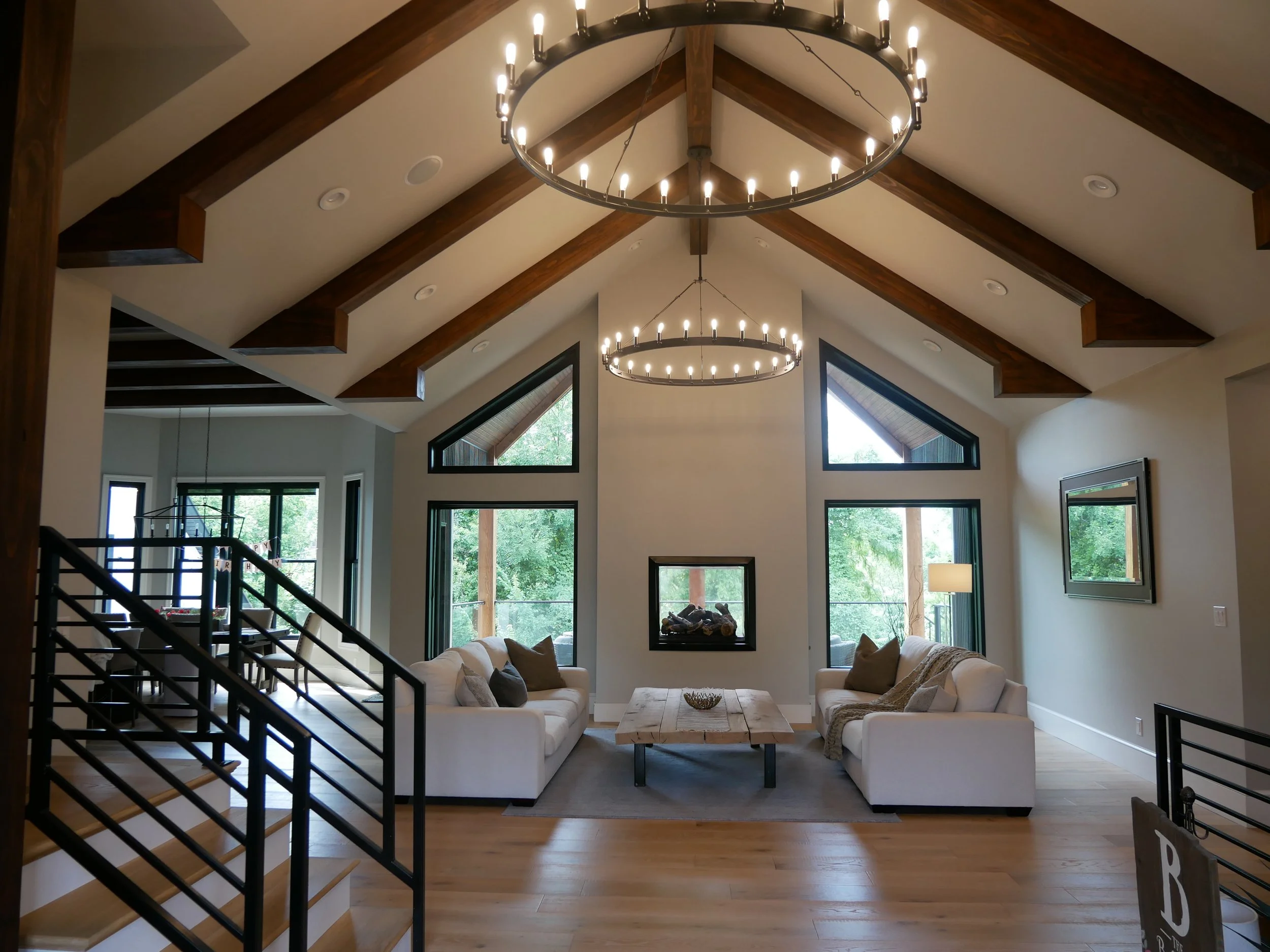 Living room with high vaulted ceiling, wooden beams, circular chandelier, large windows, white sofas, and a wooden coffee table.