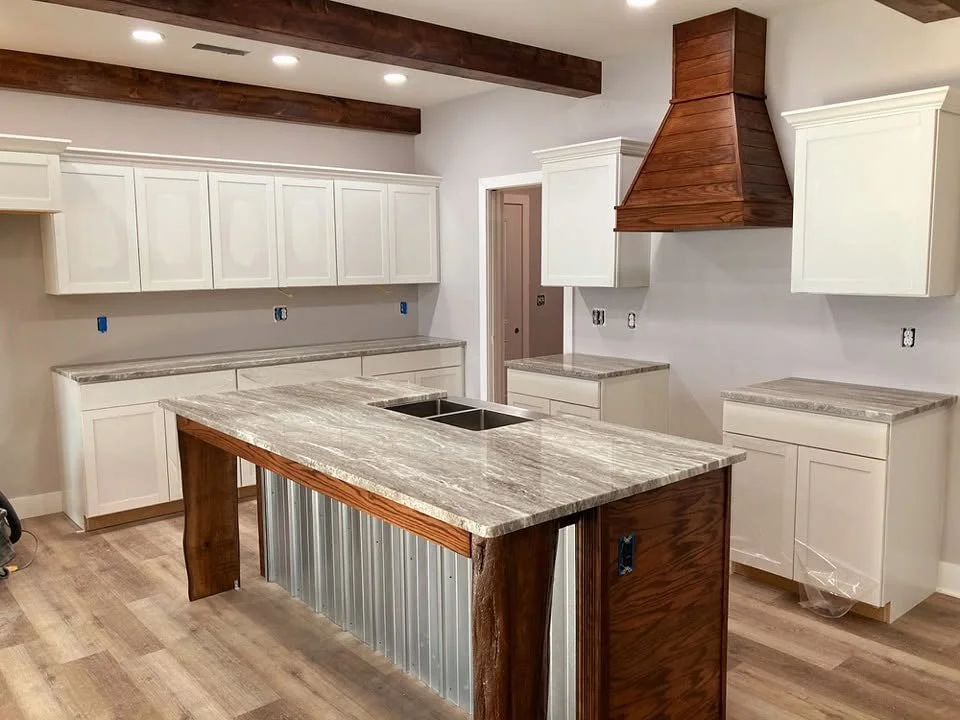 A kitchen under construction with white cabinets, a wooden range hood, a kitchen island with a stone countertop, and wooden beams on the ceiling.