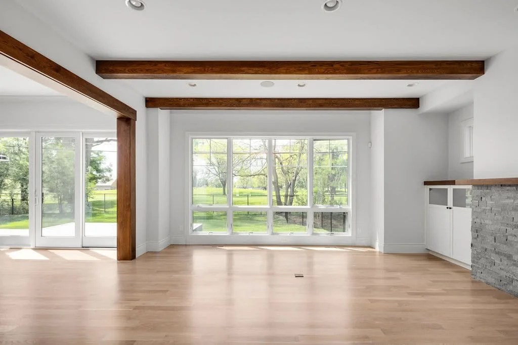 Empty living room with large windows, wooden beams on ceiling, hardwood floors, and a sliding glass door leading outside.