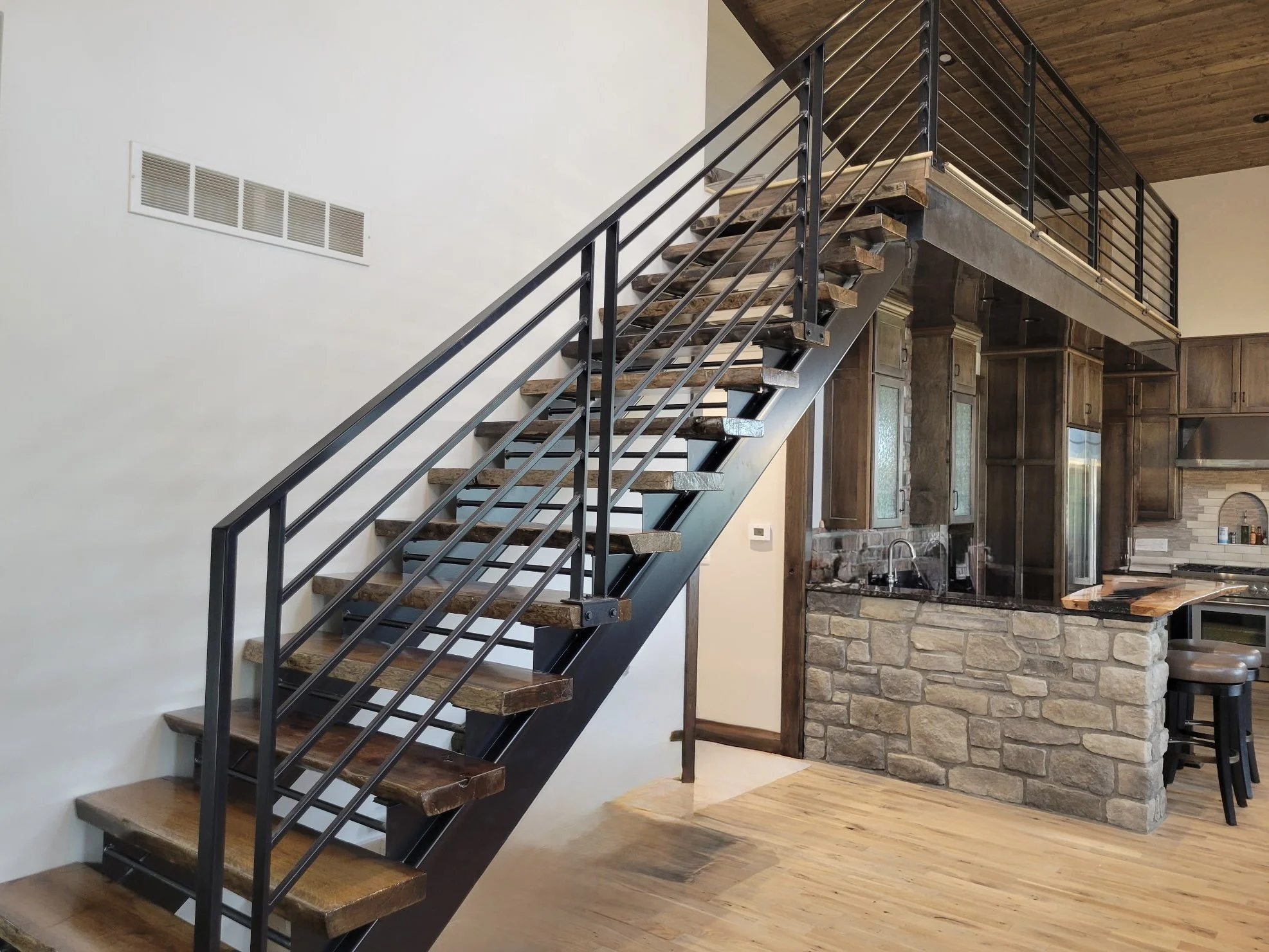 Interior view of a modern staircase with wooden treads, black metal railings, and an open space, adjacent to a kitchen with wooden cabinets and a stone wall. A vent is visible on the white wall to the left.