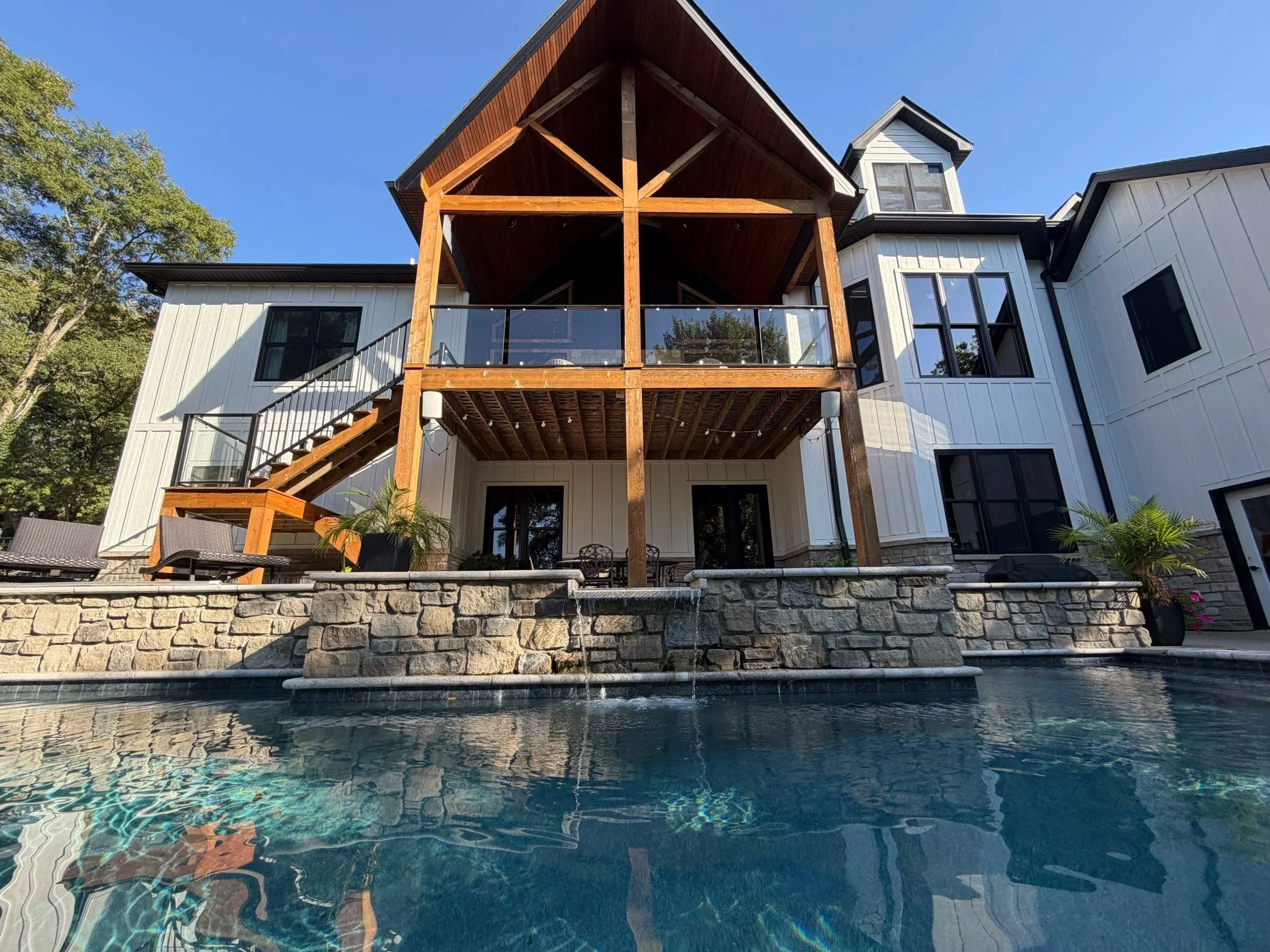 A modern house with a large deck and glass railing, a staircase leading to the yard, and an in-ground swimming pool in the foreground. The house features white siding, large windows, and a wooden overhang supported by log columns. Trees surround the property, and the sky is clear and blue.