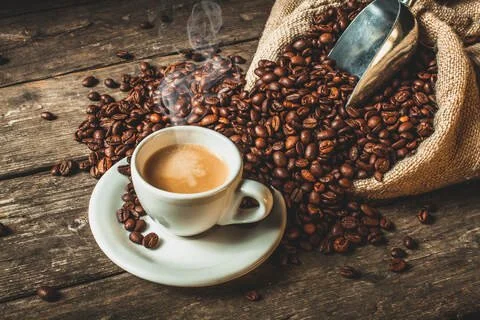 Steaming cup of coffee on a saucer surrounded by roasted coffee beans and a burlap sack on a rustic wooden table
