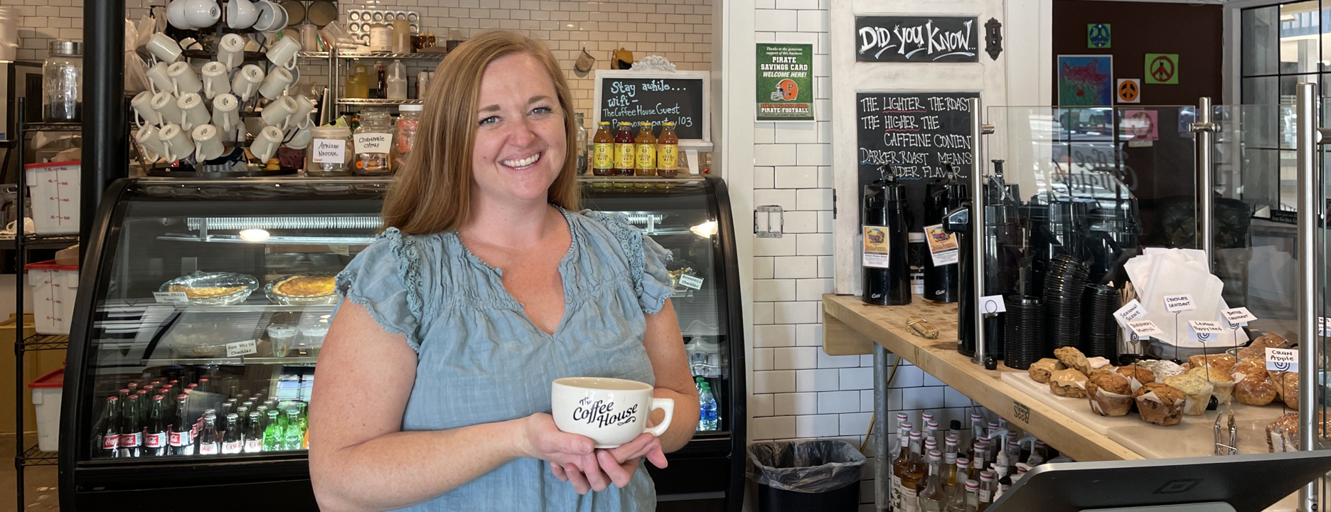 Smiling barista holding a Coffee House mug inside a cozy cafe with pastries, coffee cups, and display cases.