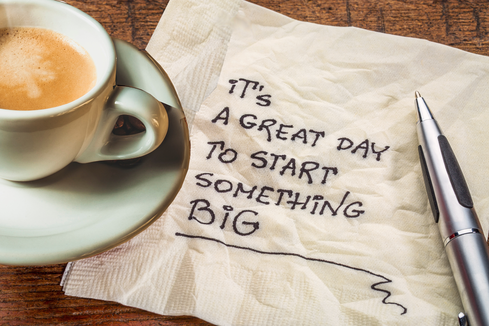 Coffee cup, pen, and napkin with handwritten motivational message, “It’s a great day to start something big,” on a wooden table.