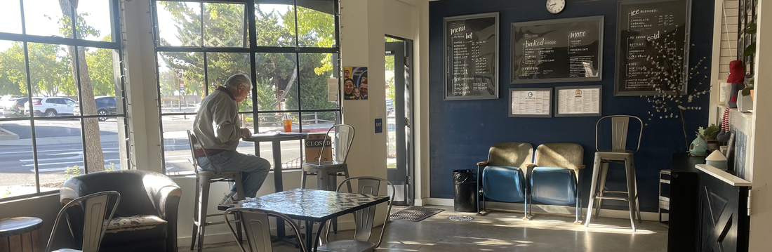Sunlit coffee shop interior with a customer seated at a window counter, tables and metal chairs, lounge seating, and menu boards on a dark blue wall