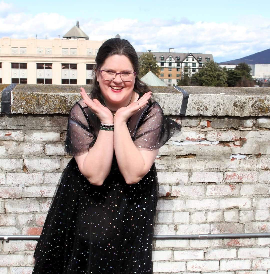 A woman with glasses and dark hair smiling and posing with hands under her chin in front of a brick wall with buildings and a mountain in the background.