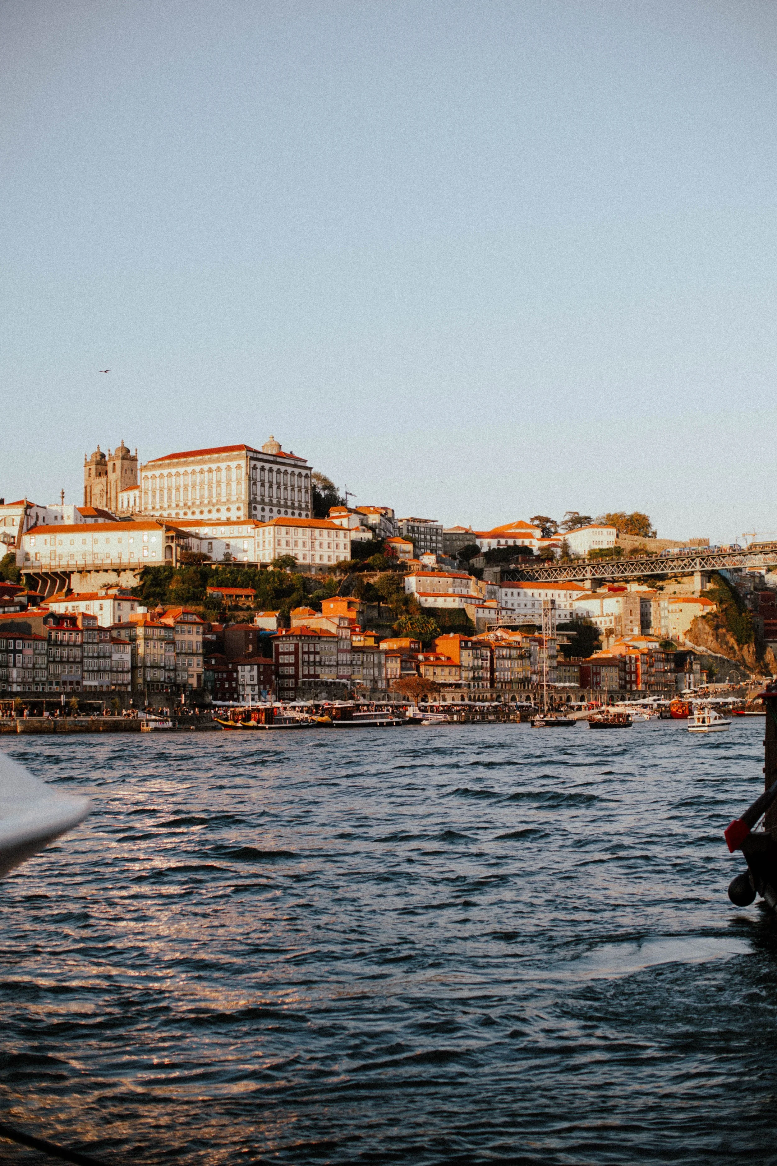 Vista de uma cidade histórica com prédios antigos e uma igreja, ao fundo, localizada na encosta, vista de um rio ou corpo de água com barcos. Cenário ao entardecer com céu claro e luz suave.