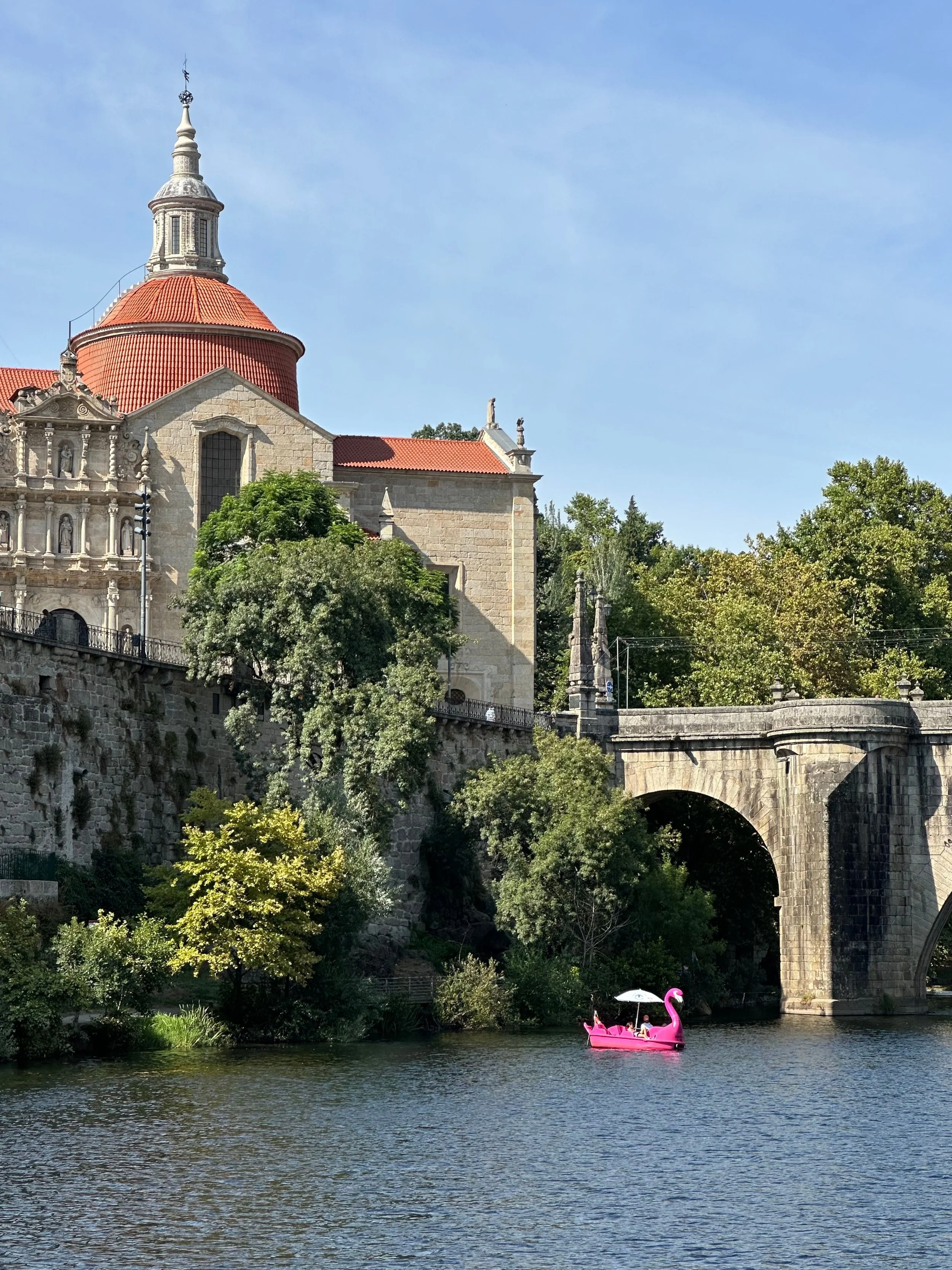 Catedral de pedra com teto vermelho, situada acima de um rio. No rio, um barco rosa em forma de cisne com duas pessoas. Árvores verdes ao redor e um céu azul claro.