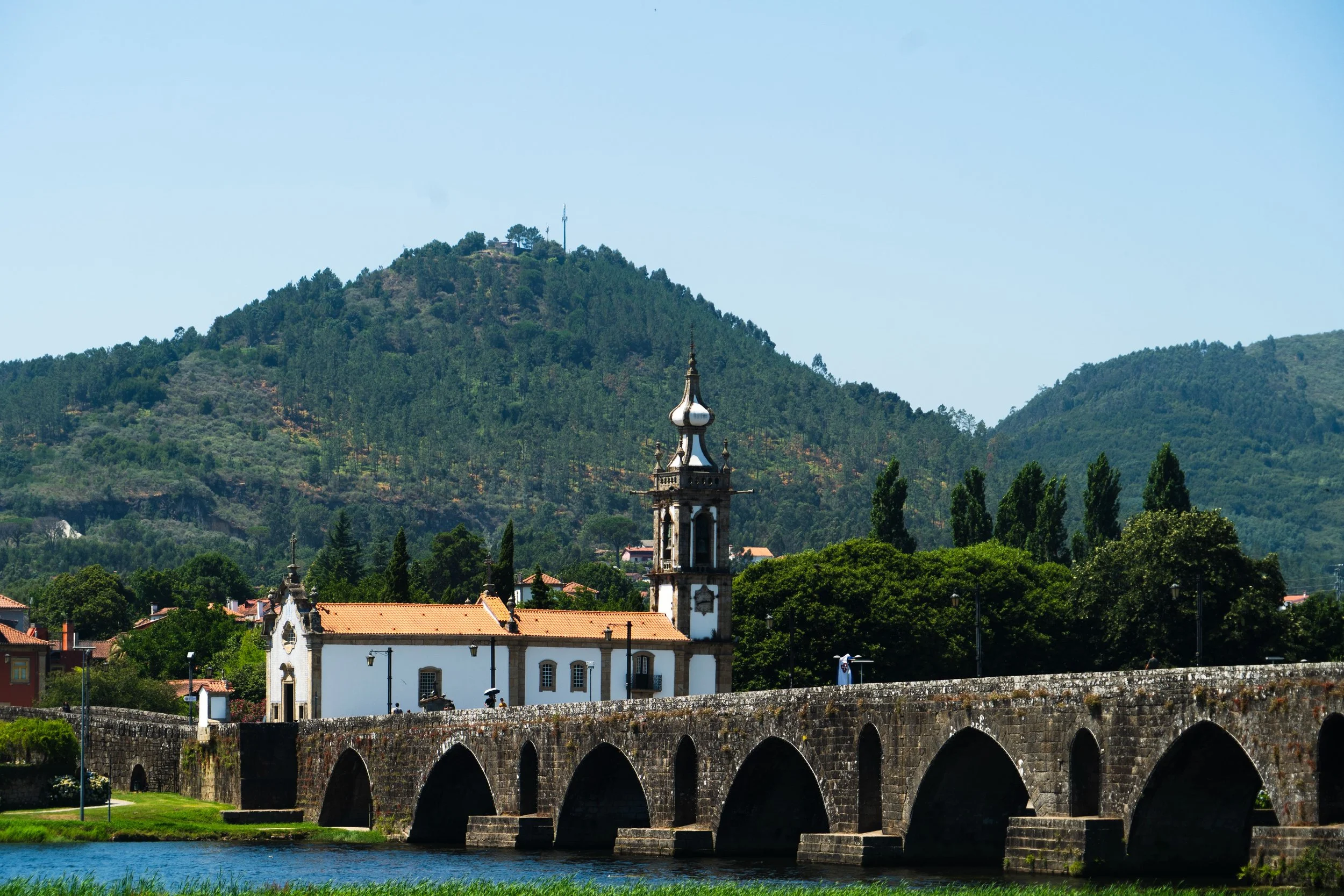 Paisagem com montanhas cobertas de árvores, uma igreja com torre ao centro, um rio na frente, e um arco de pedra na margem do rio.