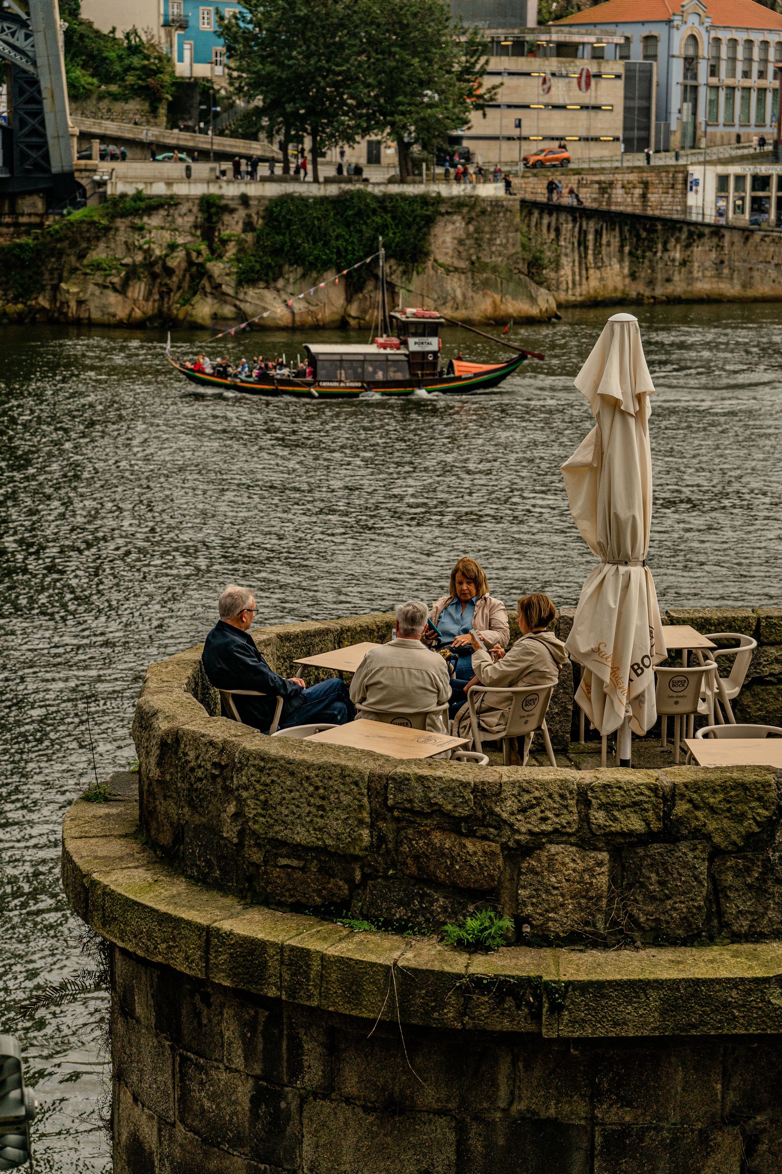 Pessoas sentadas em uma praça de pedra à beira de um rio, com um barco cheio de turistas ao fundo e edifícios coloridos na margem oposta.