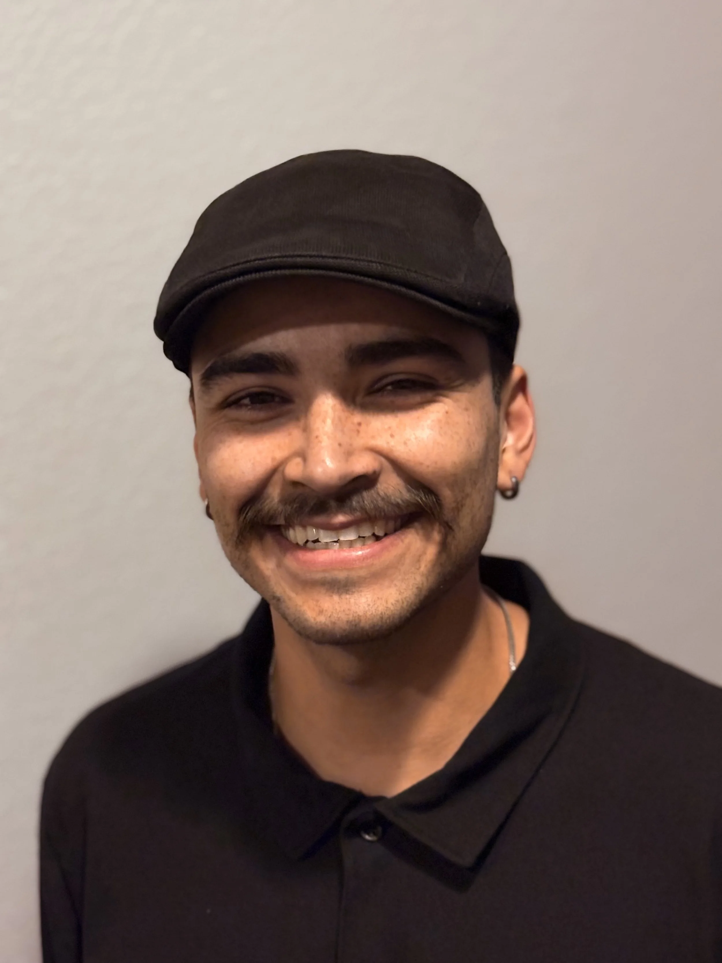Close-up photo of a smiling young man wearing a black cap and a black button-up shirt, standing against a plain light-colored wall.