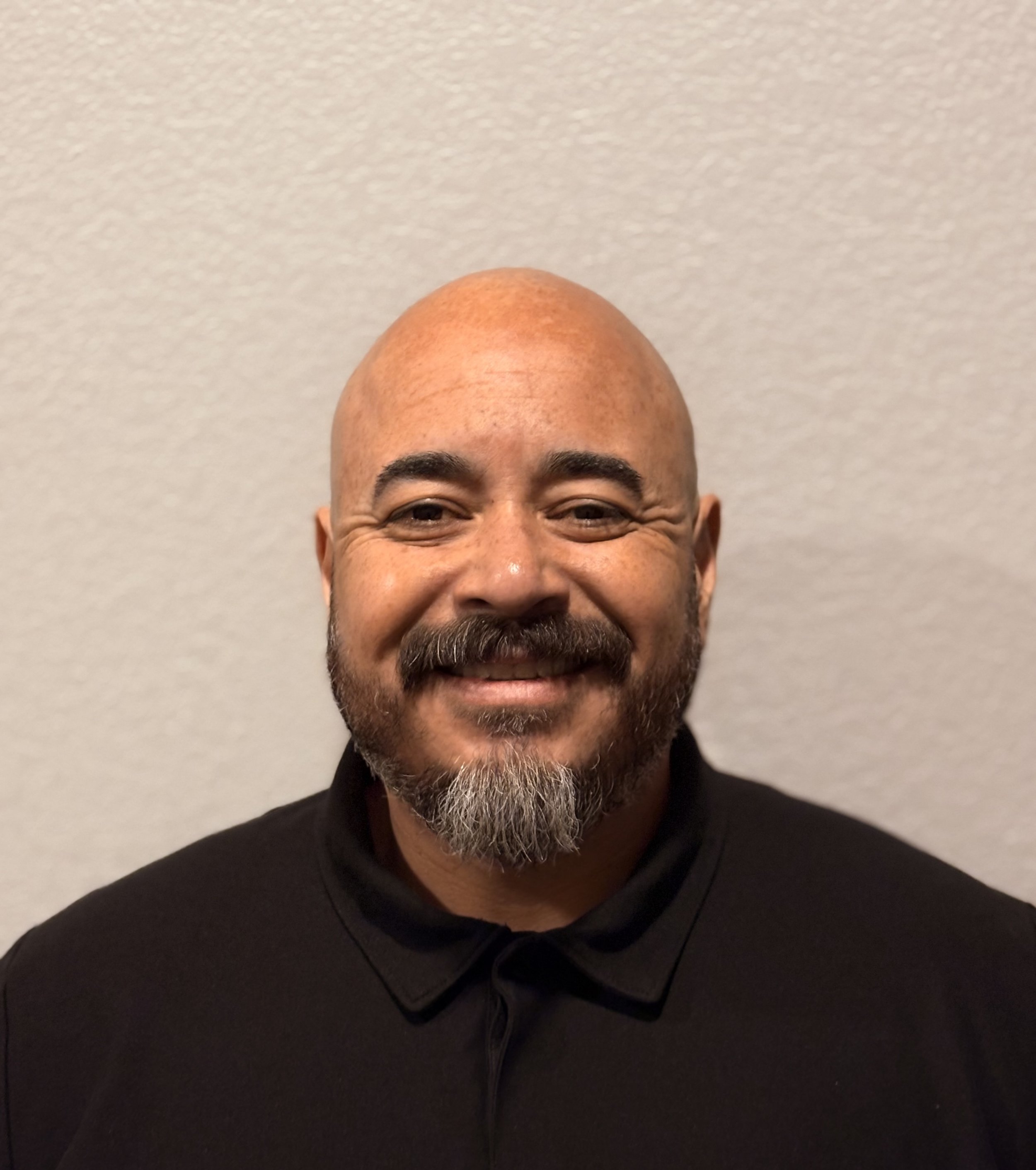 A person with a shaved head, smiling, with a beard and mustache, wearing a black hoodie, standing against a plain gray wall.