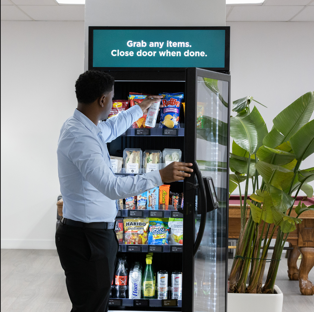 A man wearing a white dress shirt and black pants is reaching into a vending machine to grab snacks. The vending machine has various snack items like chips and candy, and a digital sign above it reads, 'Grab any items. Close door when done.' in white text on a teal background. There is a large green potted plant to the right of the vending machine.