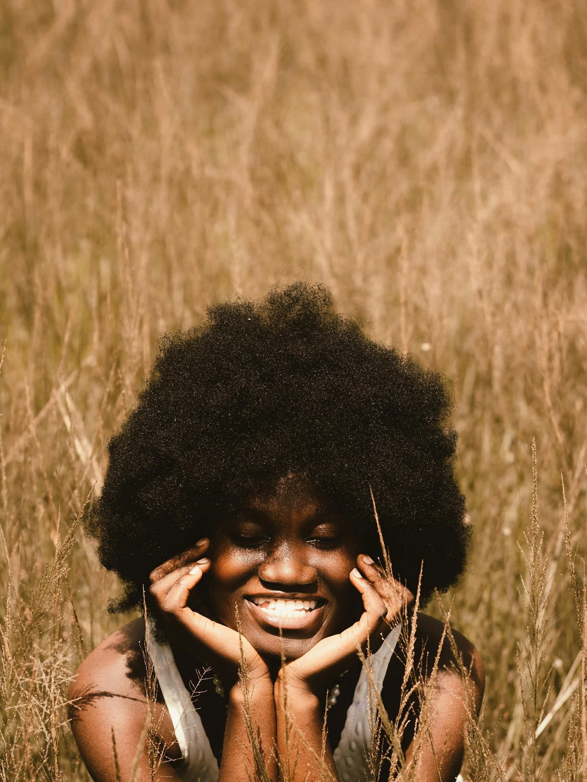 A woman with curly hair smiling and touching her face while sitting in a field of tall, dry grass.
