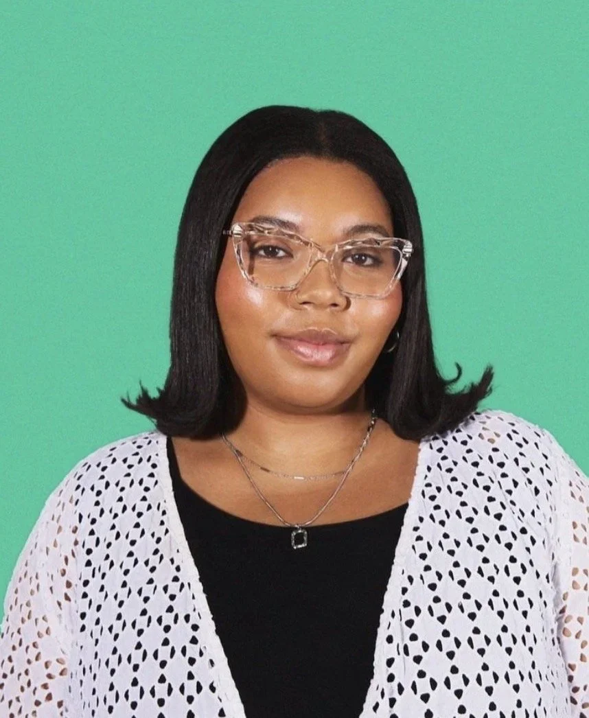 Nia, the therapist. A Black woman with medium brown skin, black hair, wearing clear glasses, a black top, a white lace cardigan, and layered necklaces, standing against a green background.