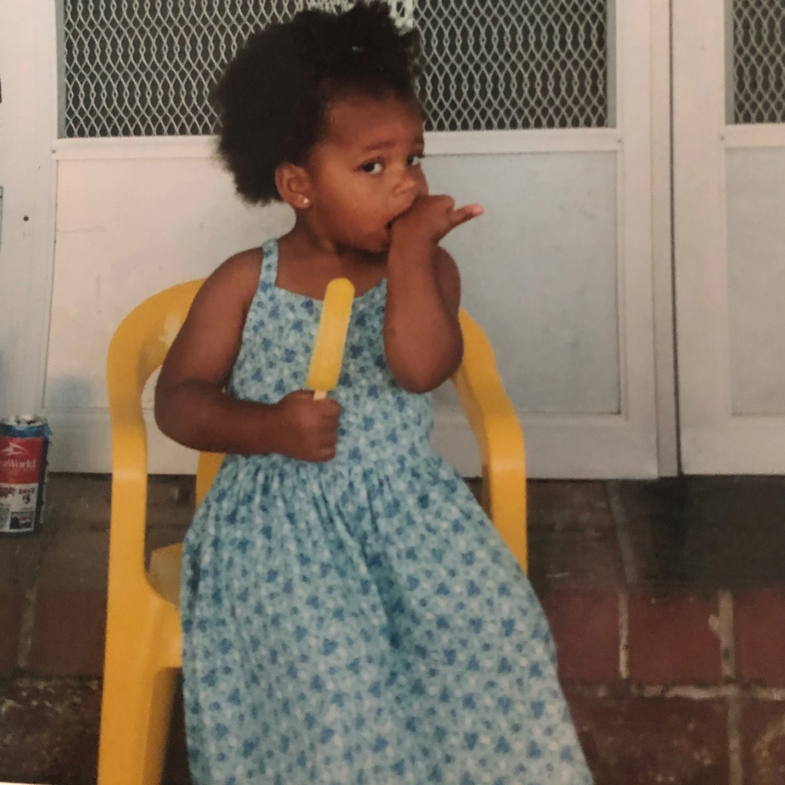 A young picture of Nia with curly hair fro, wearing earrings and a blue floral dress, sitting in a yellow high chair, biting her thumb and eating yellow popsicle.