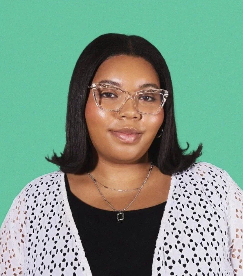 Nia, the therapist. A Black woman with medium brown skin, black hair, wearing clear glasses, a black top, a white lace cardigan, and layered necklaces, standing against a green background.