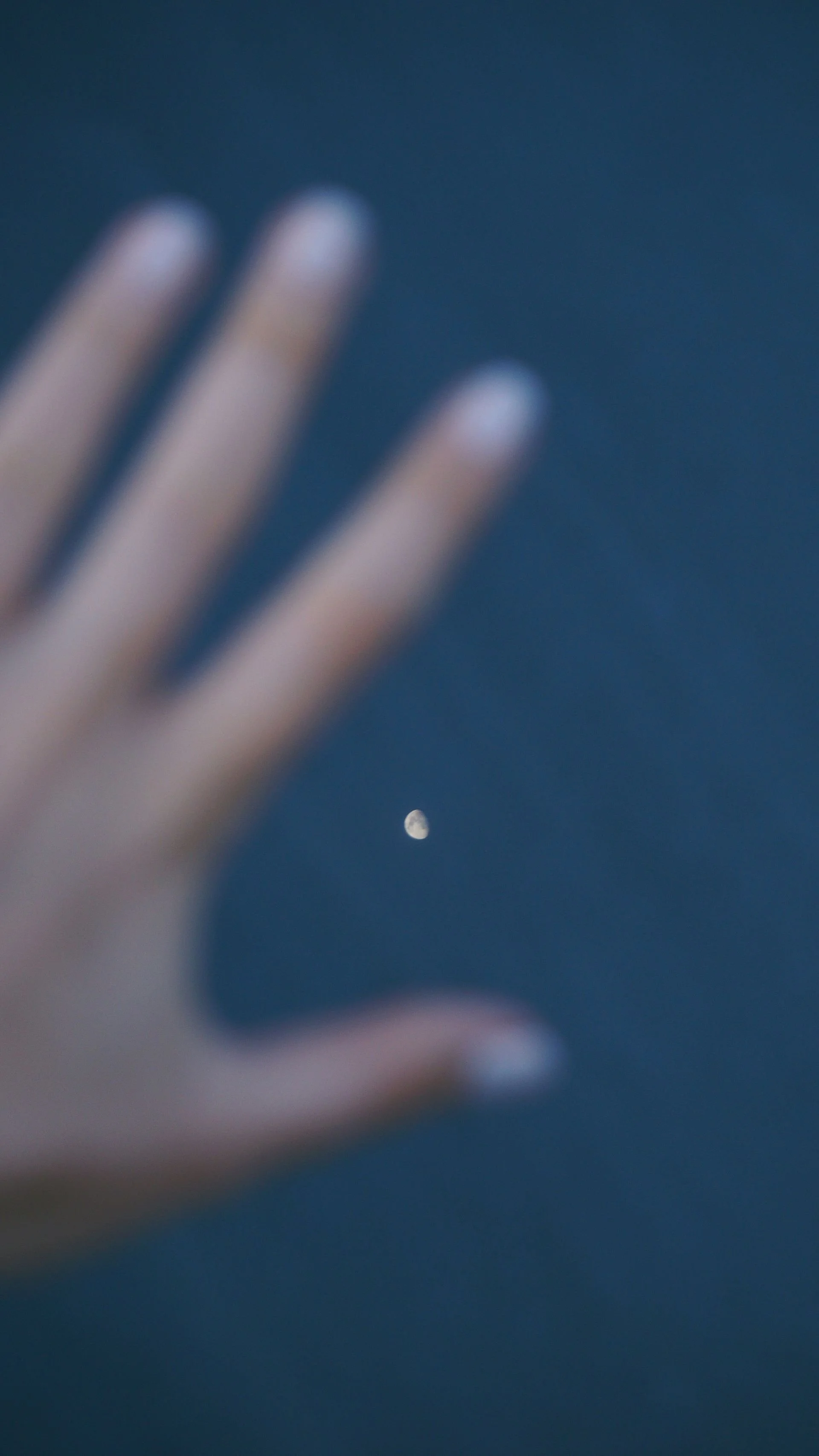 Close-up of a person's hand with fingers spread, with the moon visible in the night sky in the background.