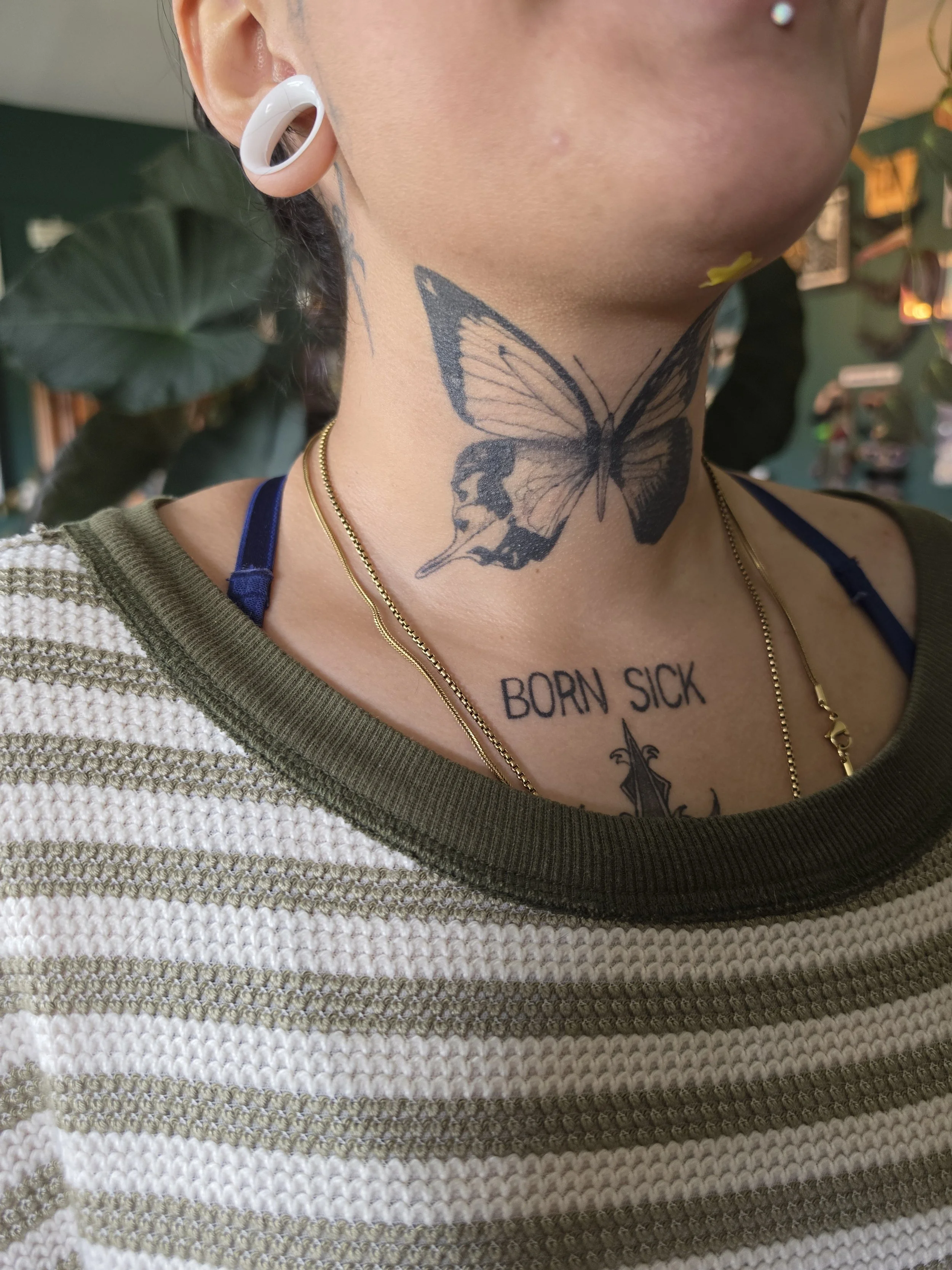 Close-up of a person's neck and part of their face, showing a butterfly tattoo on the neck and the words 'BORN SICK' tattooed on the chest. The person has a white earbud in the ear and layered necklaces.