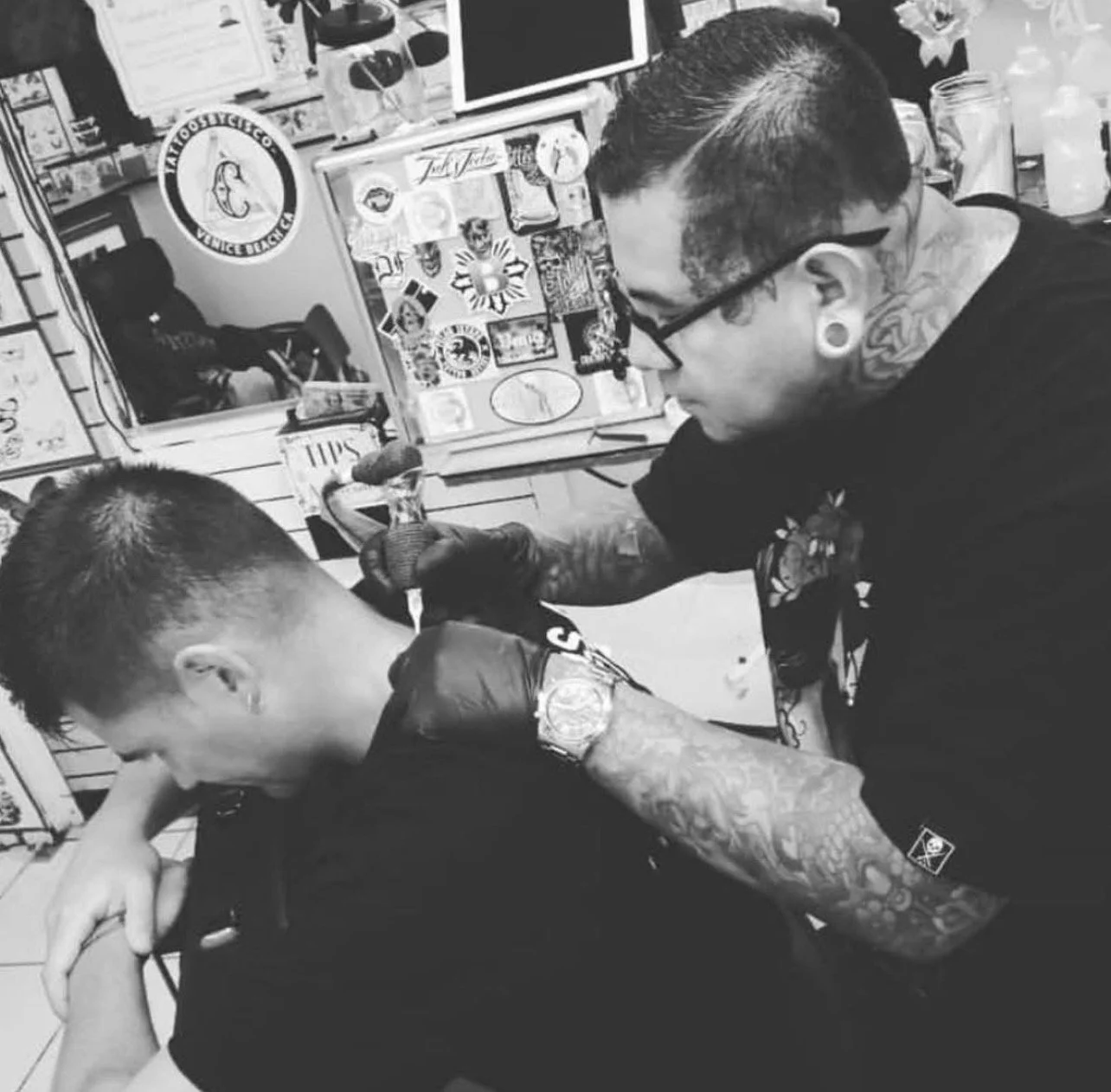 Tattoo artist tattooing a person's arm inside tattoo shop with various stickers and posters on walls.