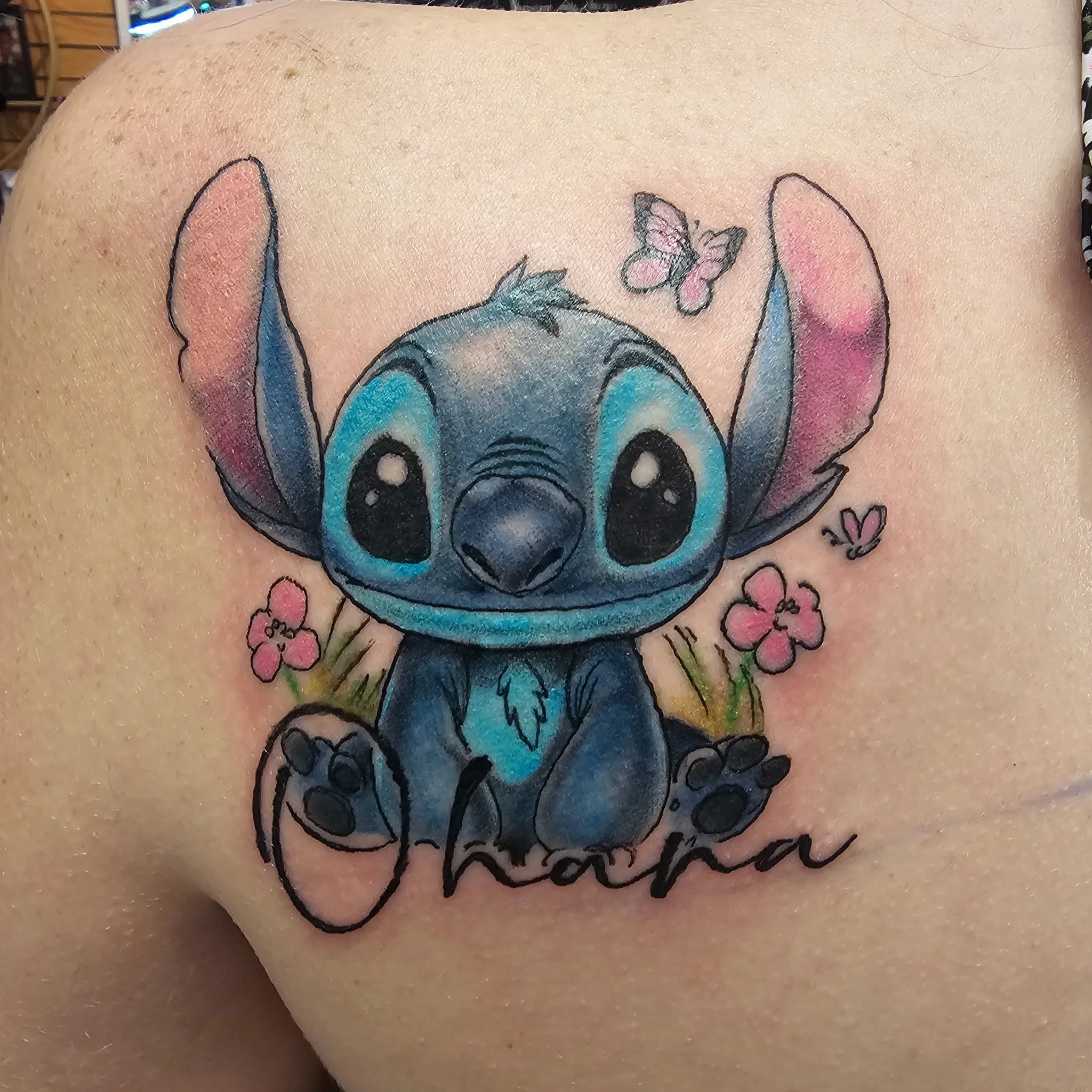 Tattoo of Stitch from Lilo & Stitch with pink and blue accents on a person's skin, accompanied by pink and green floral designs and cursive text.