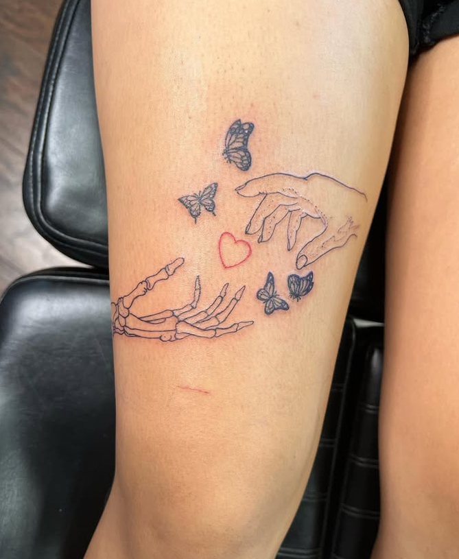 Tattoo of two human hands reaching towards each other, surrounded by butterflies and a red heart on person's thigh.