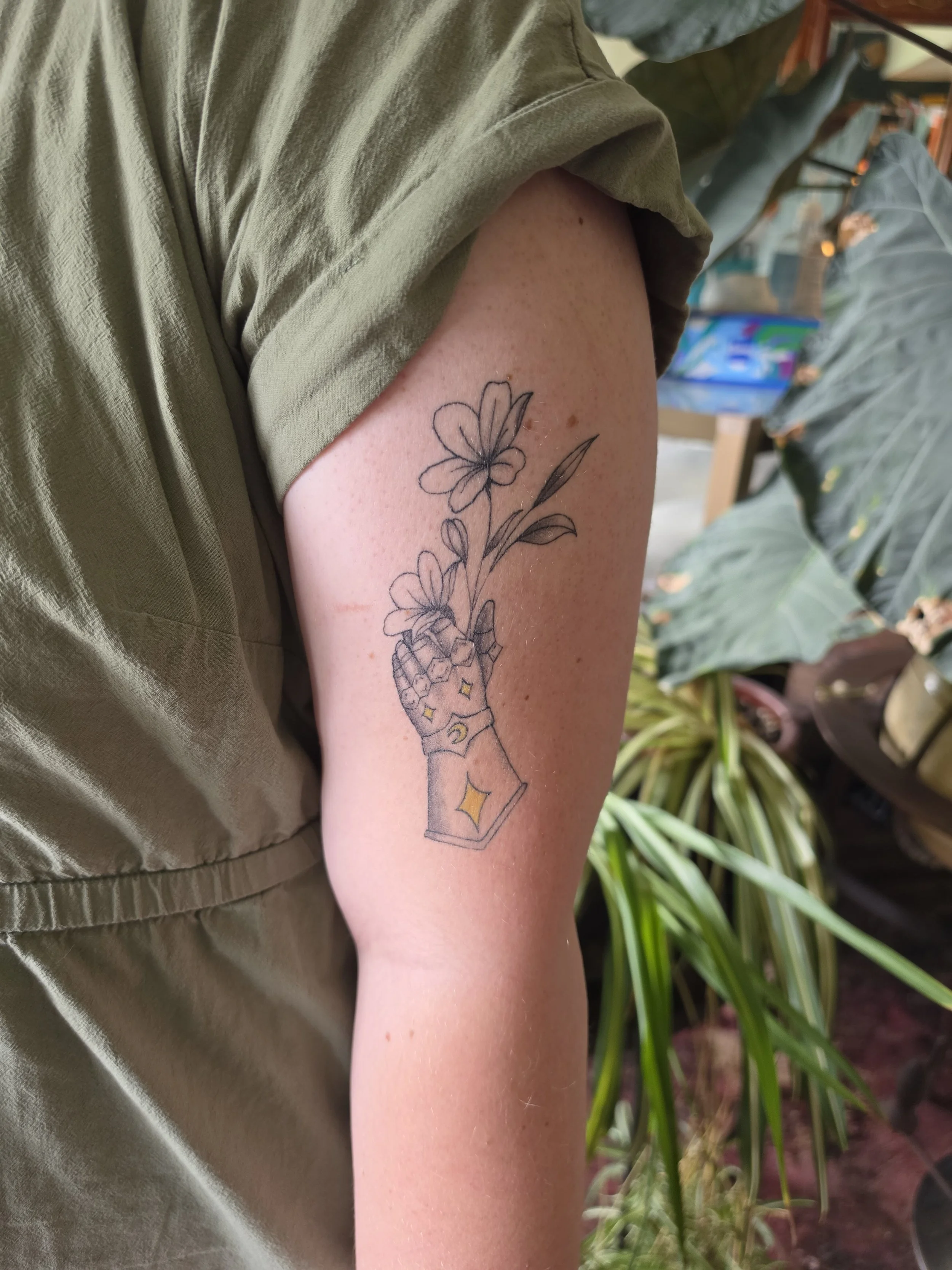 A person's upper arm with a tattoo of a flower growing out of an ice cream cone, next to other plants in the background.