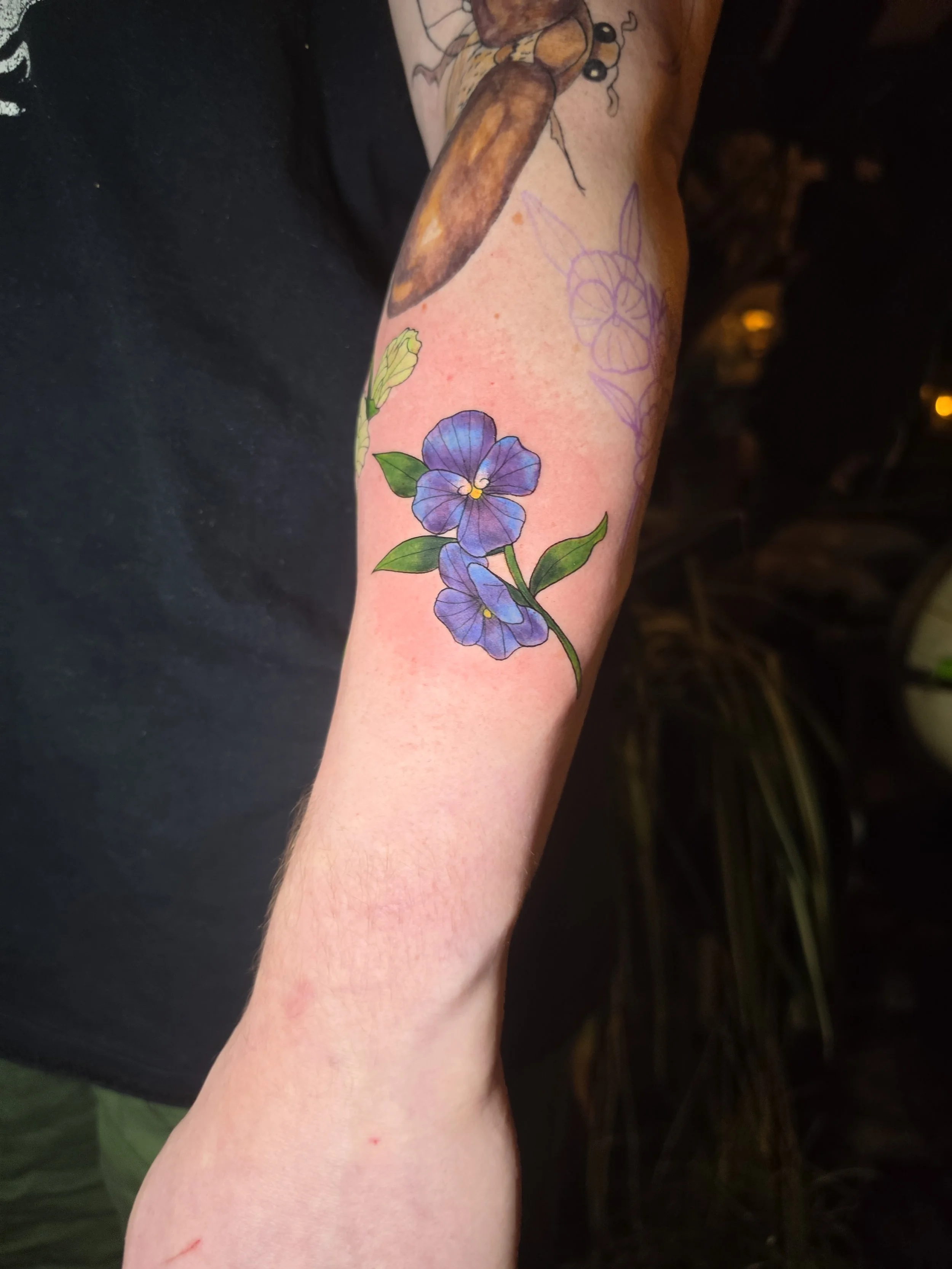 A tattooed arm with a colorful flower tattoo, showing purple flowers with green leaves and a sketch of a rabbit outline in purple ink.