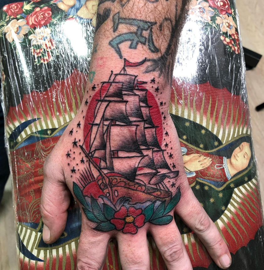 Tattoo of a sailing ship with black sails and red hull, surrounded by a red flower with green leaves, on the person's hand and wrist.