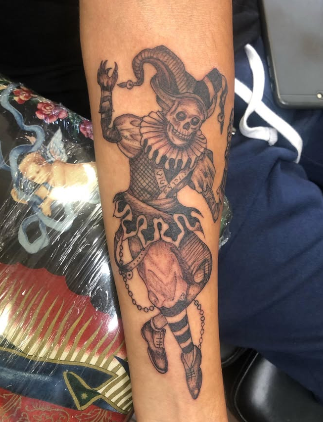 Tattoo of a clown with a skeleton face, wearing a ruffled collar and striped tights, carrying a large mask on a chain, with one arm raised and the other holding the mask.