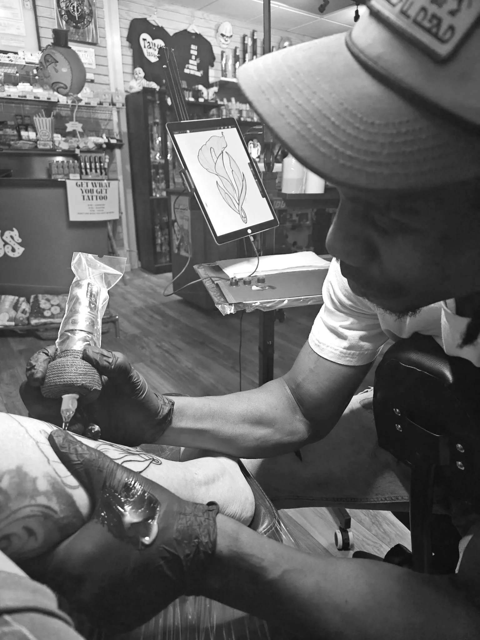 A person with tattoos on their arm receiving a tattoo in a tattoo shop. The tattoo depicts a flower on a tablet screen connected to a tattoo machine by a cord. The shop has various items on shelves, including T-shirts, masks, and decorative objects.