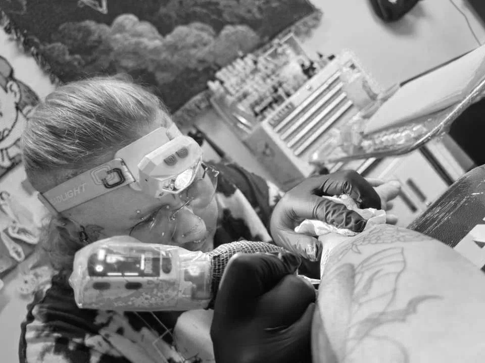 A person wearing goggles and gloves holds a tattoo machine, tattooing a design on someone's arm in a tattoo studio.
