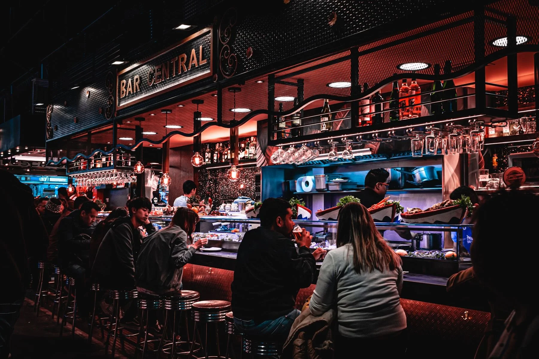 Crowded Bar Central restaurant and seafood bar with diners seated at the counter, liquor bottles, hanging glasses, and red-blue lighting