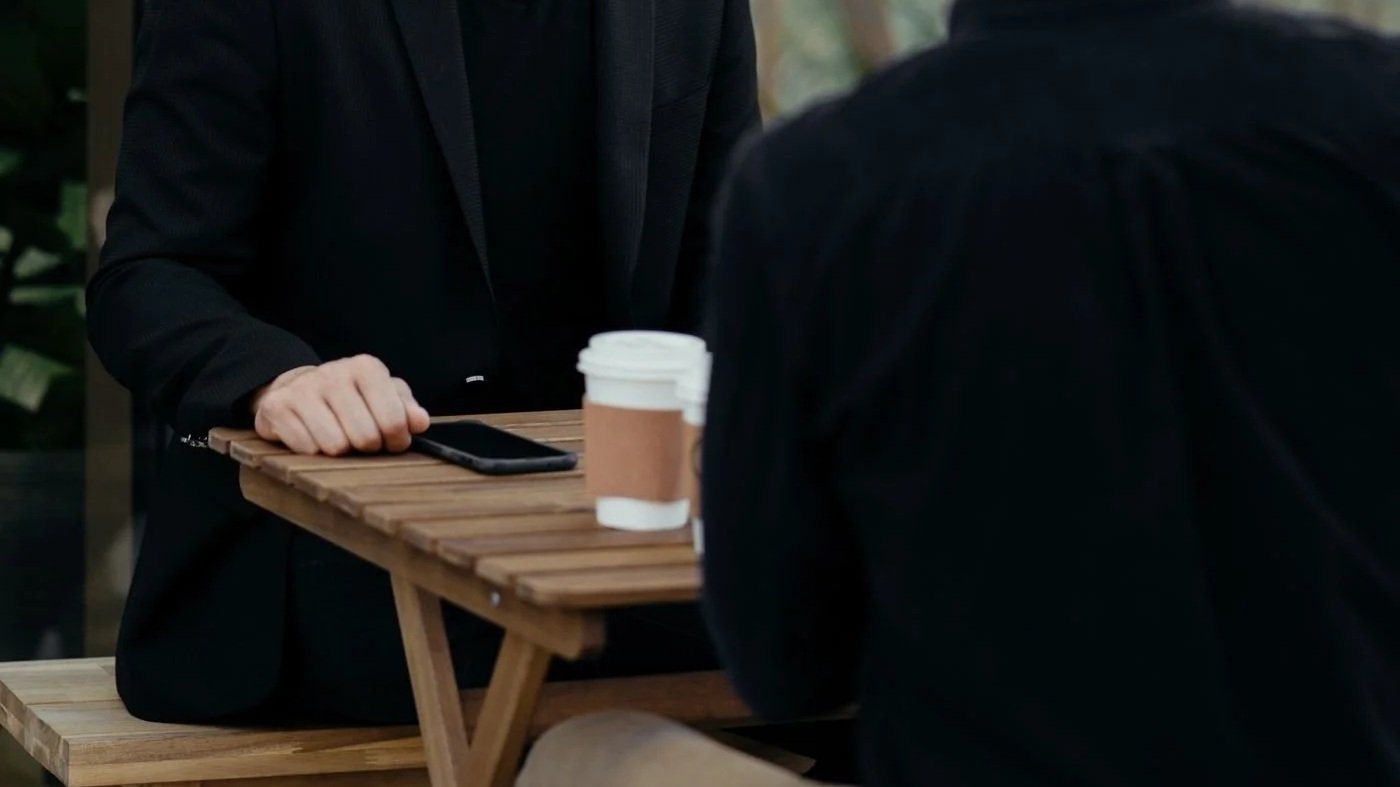 Two people in dark clothing sit at a wooden outdoor table with a smartphone and takeaway coffee cup.