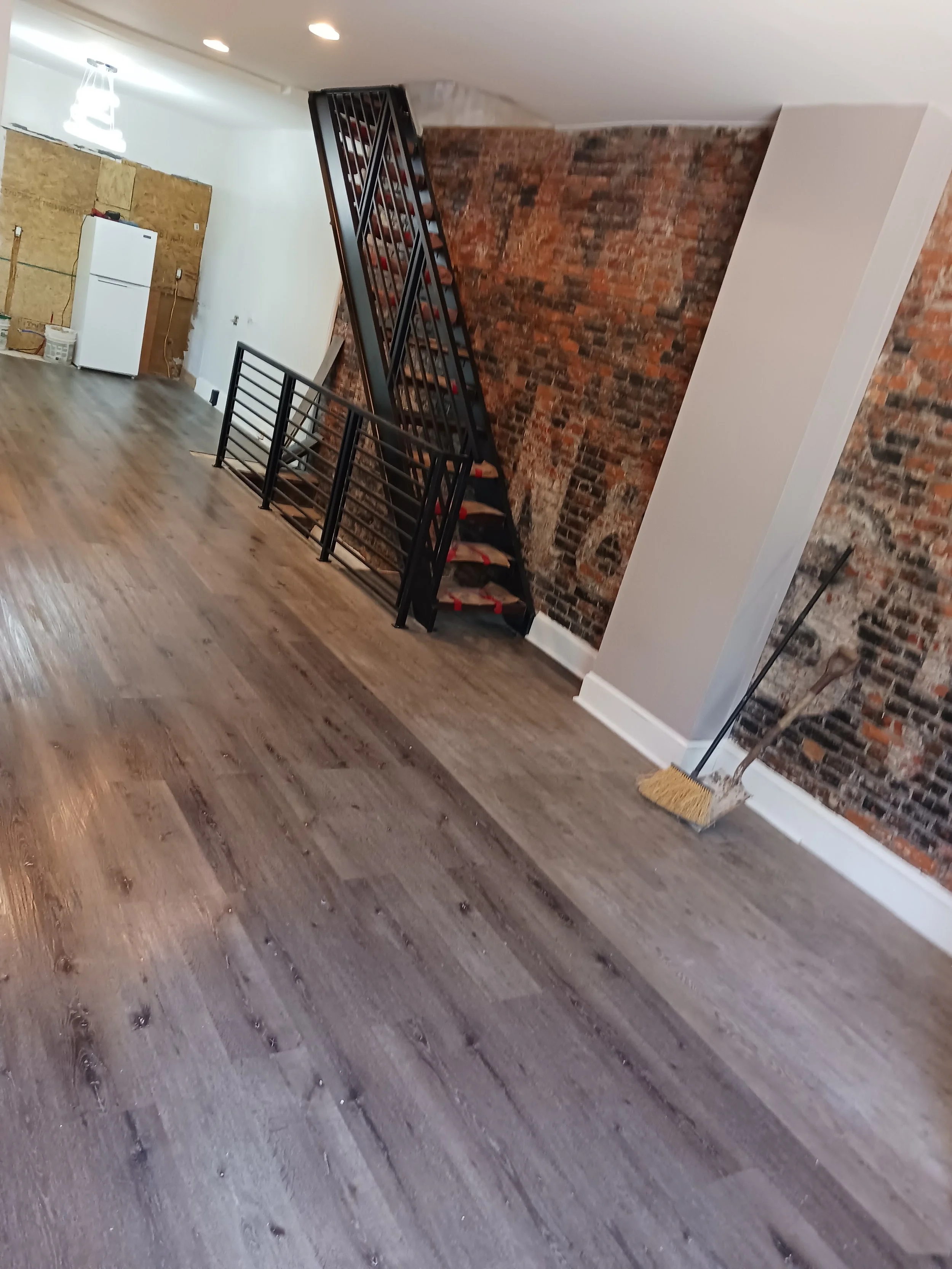 Interior of a room with wooden flooring, exposed brick walls, a white refrigerator, and construction materials.