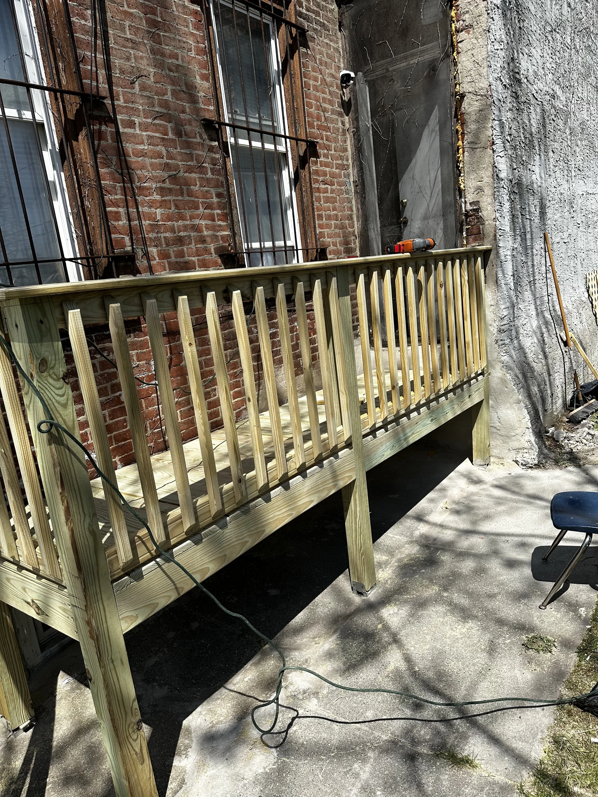 Constructed wooden deck with a rope hanging off, attached to the back of a brick building with two windows. Construction tools are on the deck, and a security camera is mounted on the building. The ground below is concrete with some grass and weeds, and a chair is in the lower right corner.