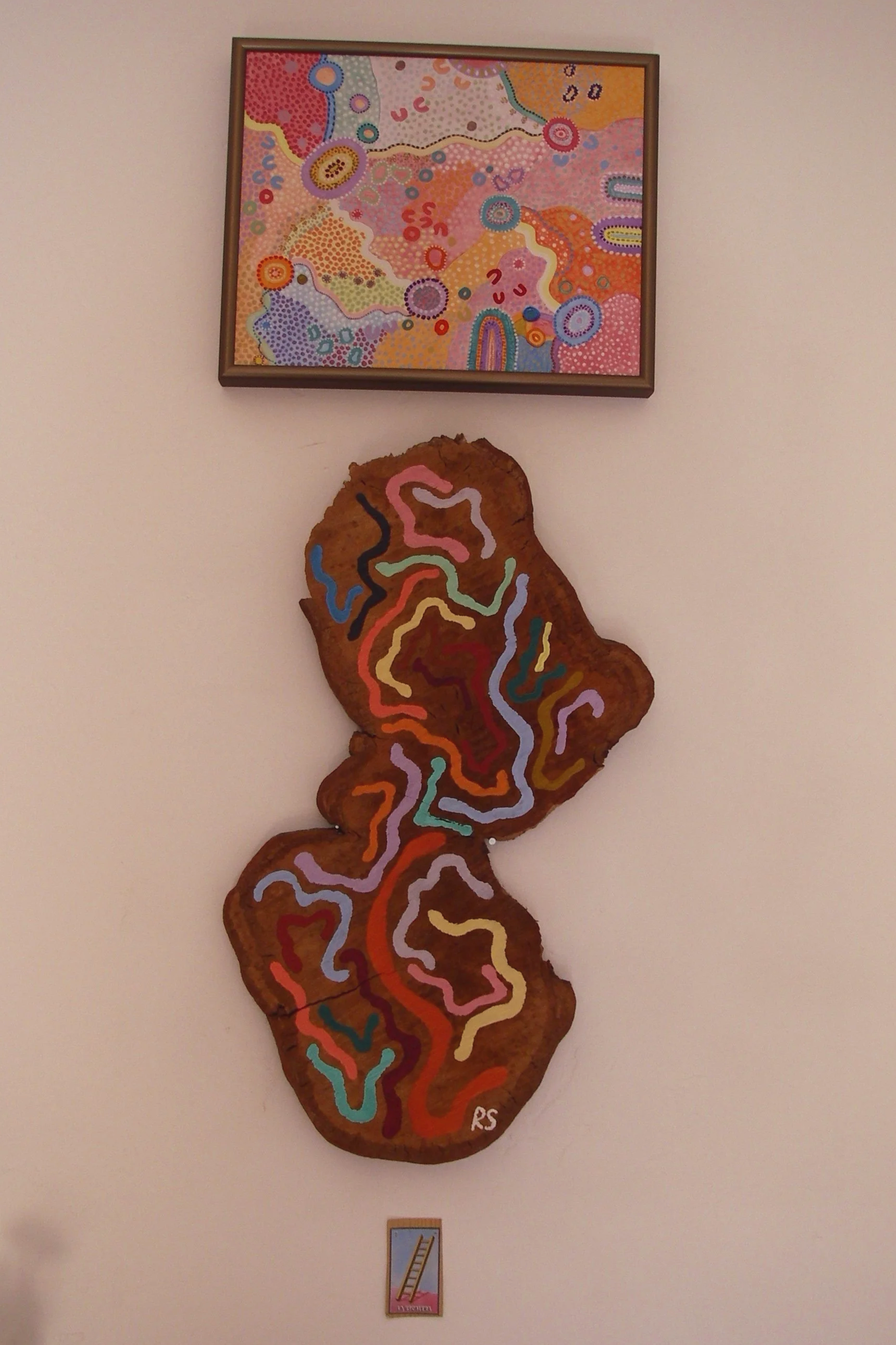 A colorful abstract painting on a wall showing various vibrant patterns and shapes, with a large wood-like cutout below it painted with a colorful abstract design, and a small framed artwork with a ladder and pink background underneath.