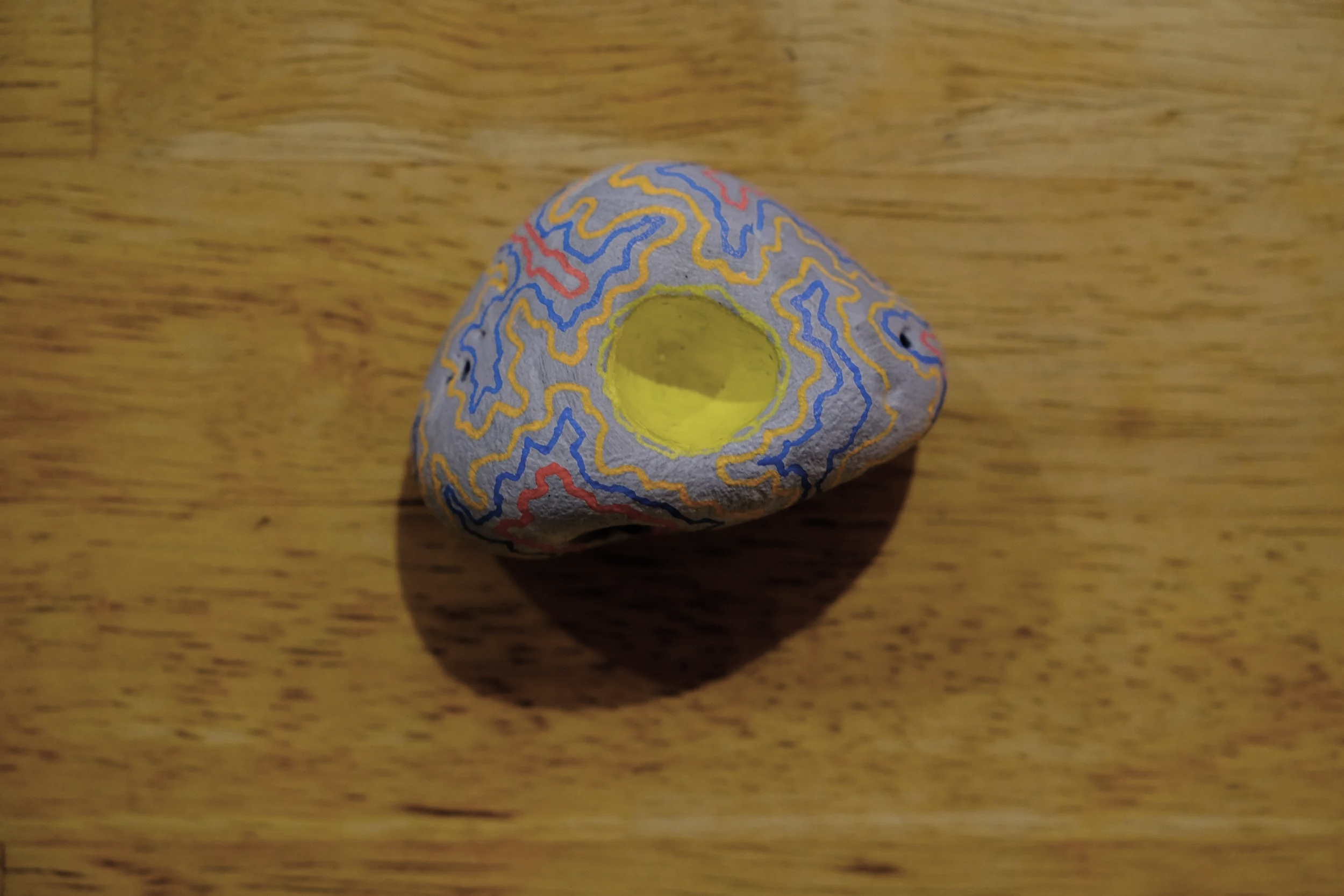 Colorful painted rock with abstract maze-like blue, red, and yellow patterns on a wooden surface.