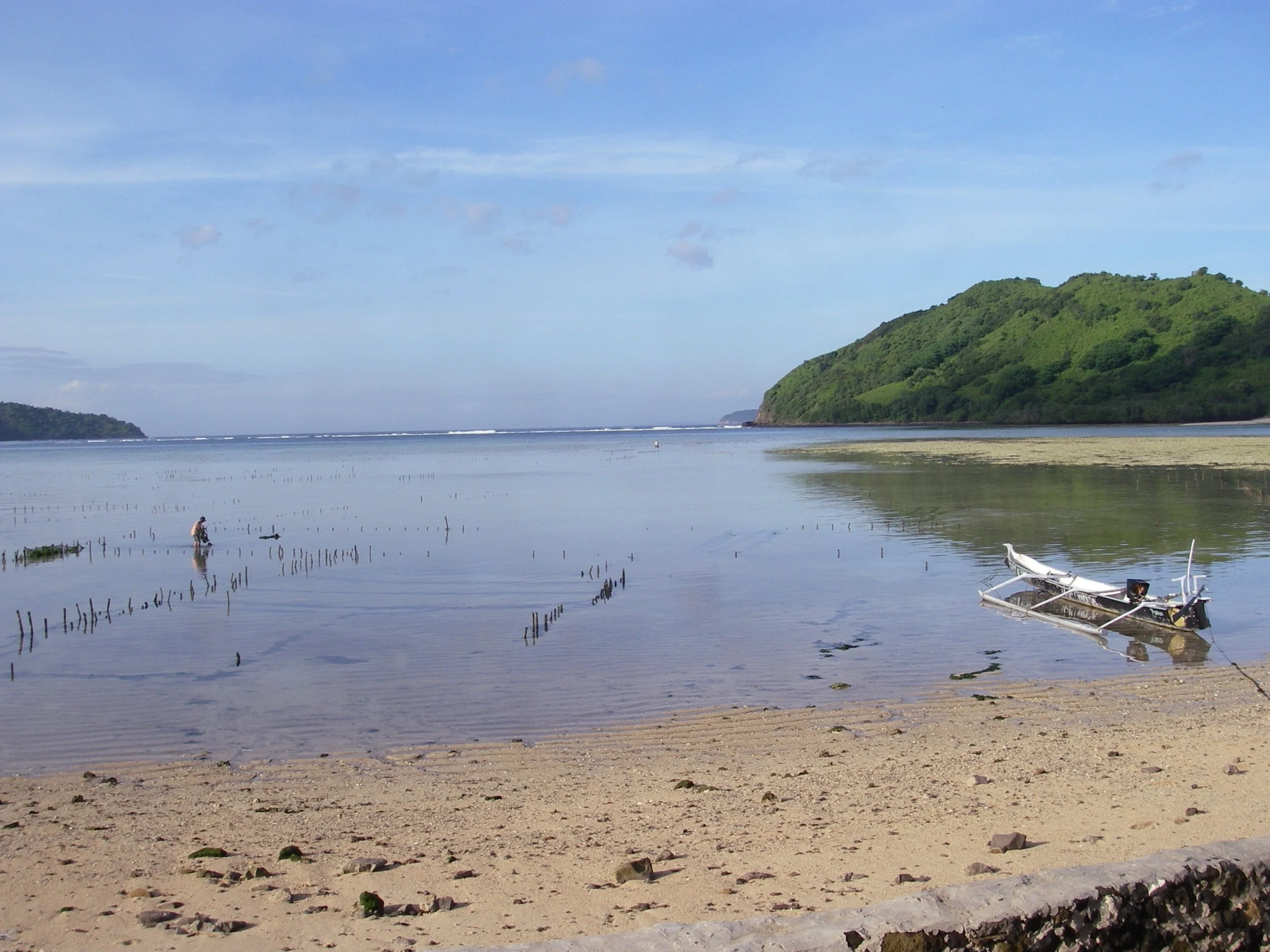 Beach with sand, a boat on the shoreline, calm water, green hill in distance, blue sky with some clouds.