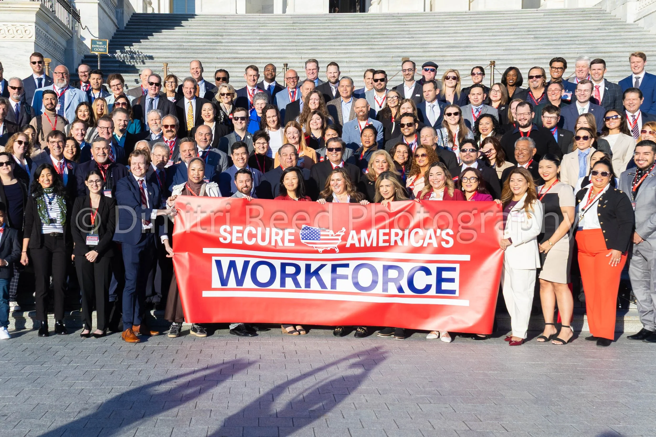 A large group of diverse people gathered on the steps of a government building, holding a red banner that reads 'Secure America’s Workforce'. The group appears to be participating in a rally or protest advocating for workforce security and immigratio