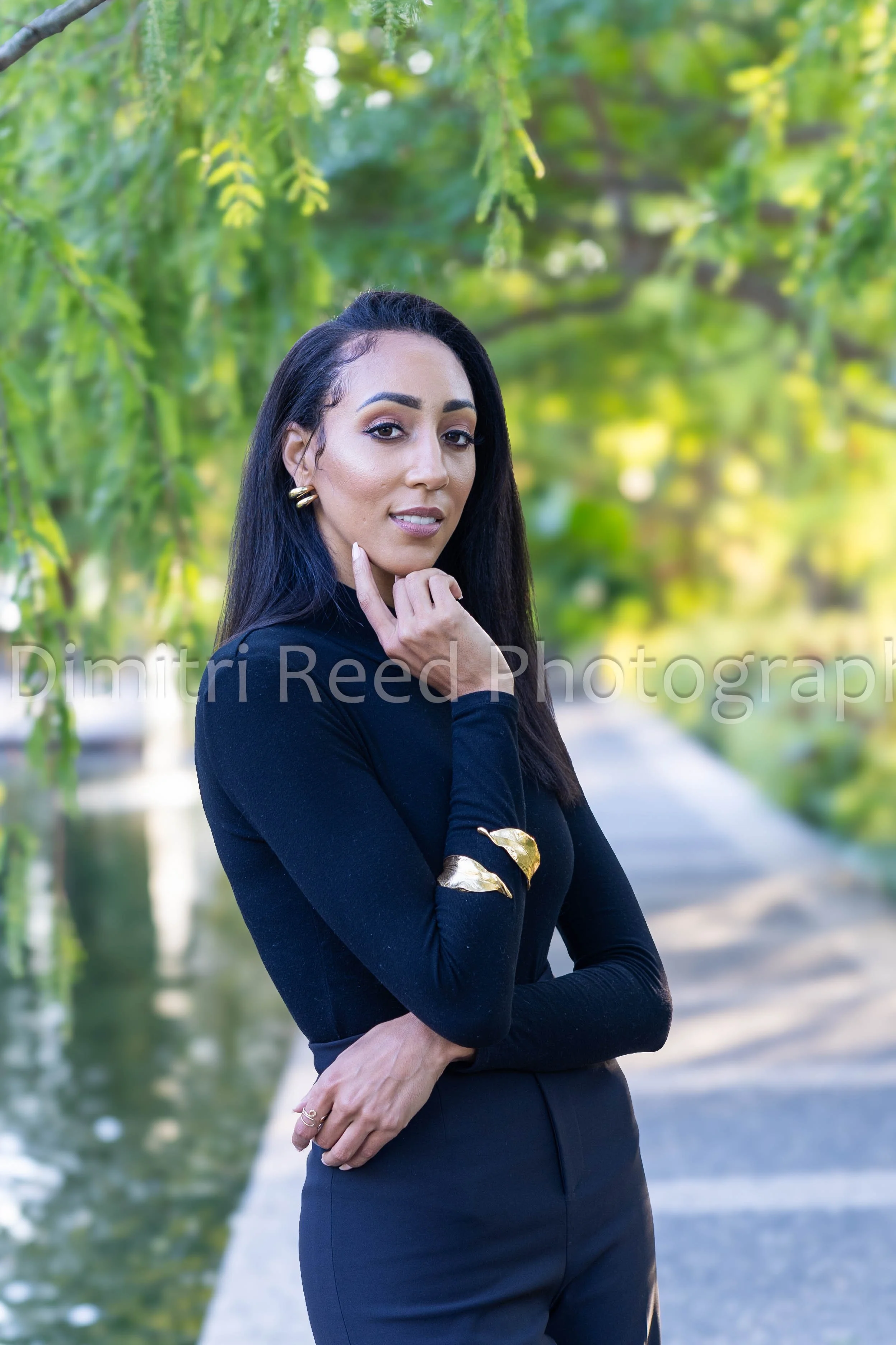 A woman in a black top and gold accessories standing outdoors near a canal with green trees in the background.