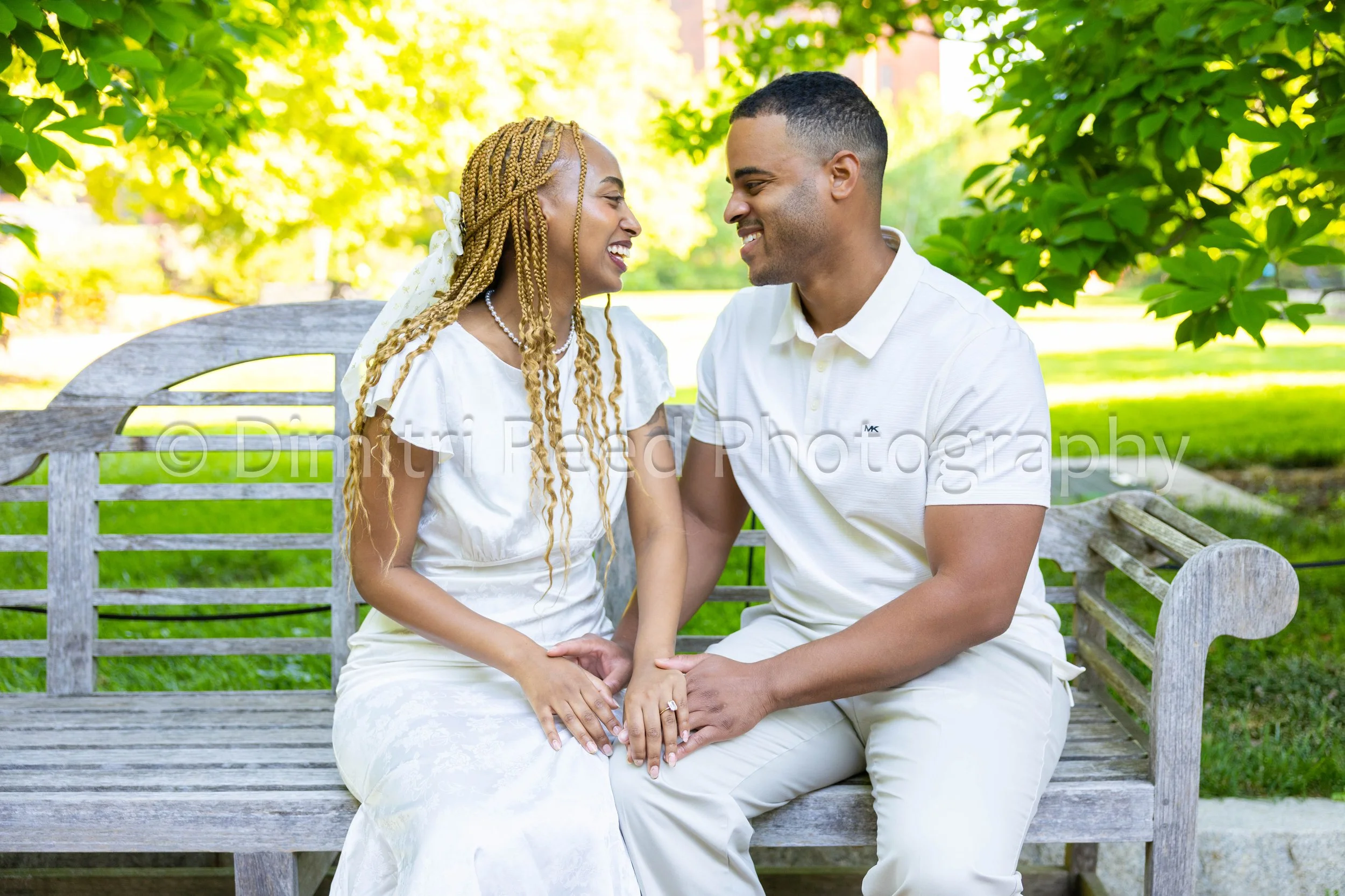A joyful couple sitting close together on a park bench, holding hands and smiling at each other. The woman has blonde braids with a white bow, wearing a white dress, and the man has short black hair and a white polo shirt. They are surrounded by lush